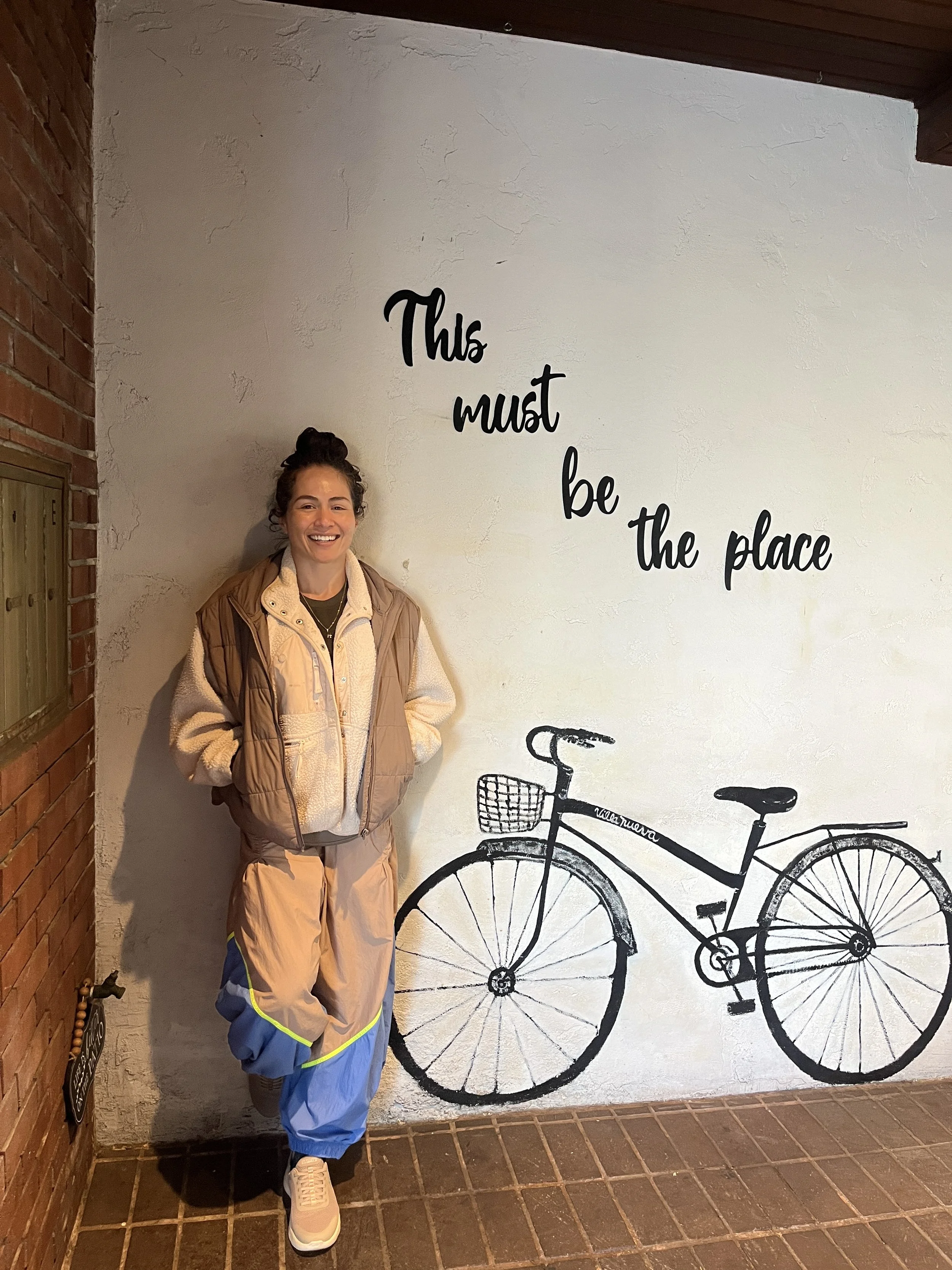 A smiling woman with curly hair standing in front of a wall with a bike illustration and the phrase "this must be the place" written on it. She is wearing a beige jacket over a cream sweater, and baggy beige and blue pants with white sneakers.