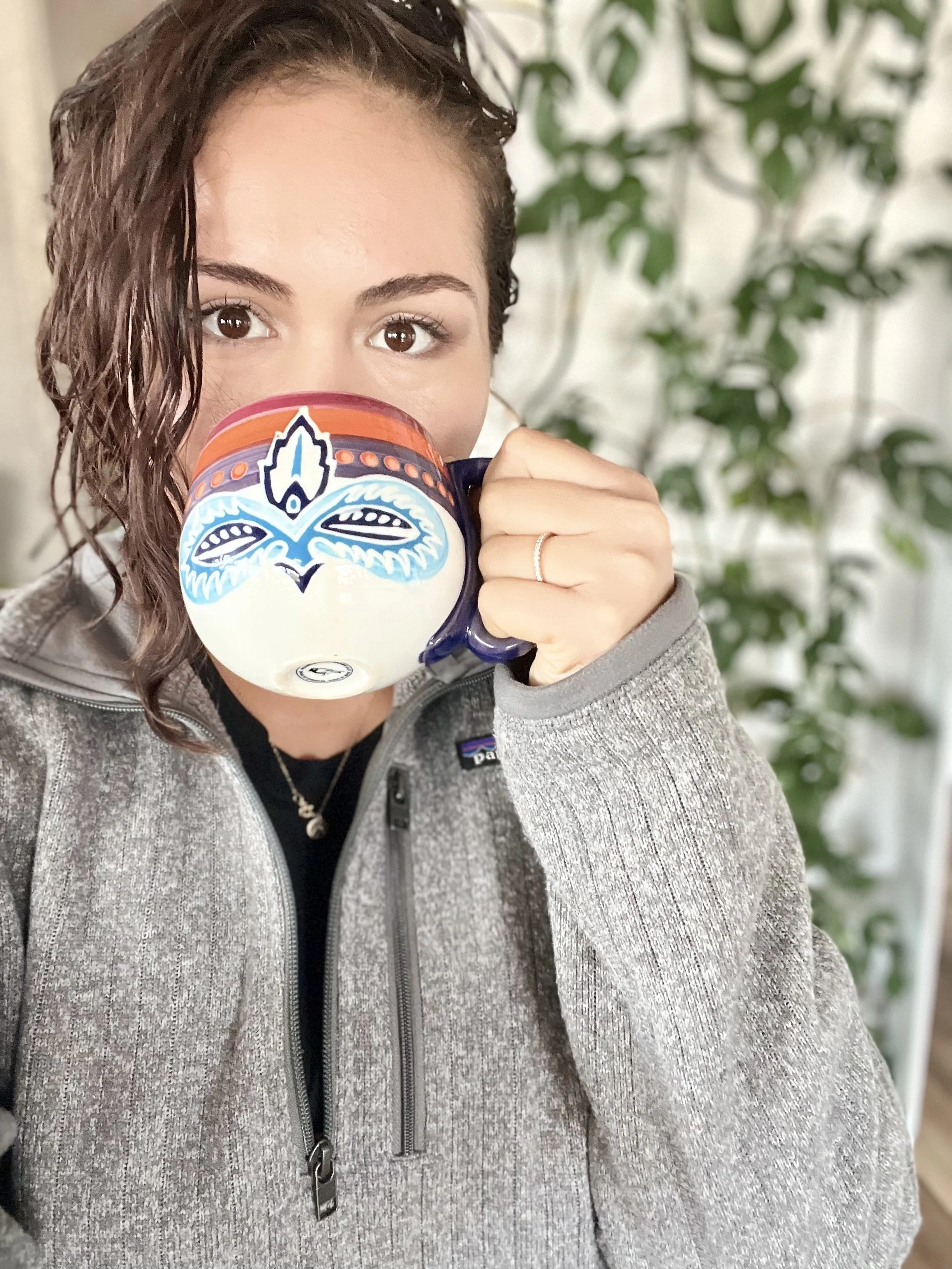 A woman with curly brown hair looking at the camera while holding a colorful mug over her face, with a background of green foliage.
