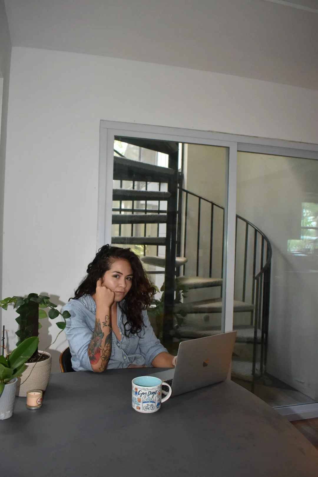A woman with curly hair, tattoos on her left arm, sitting at a dining table, looking at a laptop, with a window and spiral staircase in the background. There are potted plants, a candle, and a coffee mug on the table.