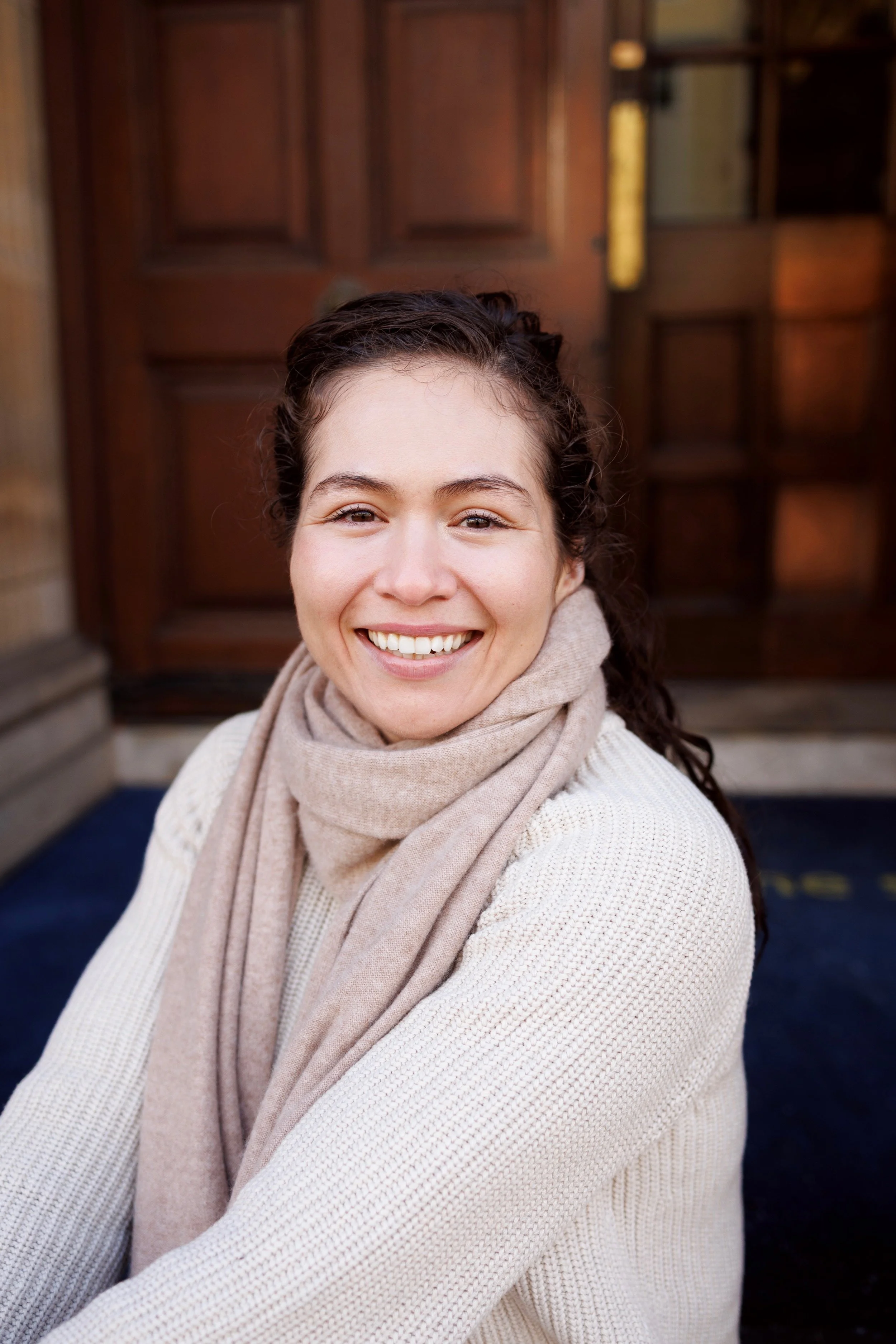 A woman smiling outdoors in front of a wooden door, wearing a beige scarf and light-colored sweater.