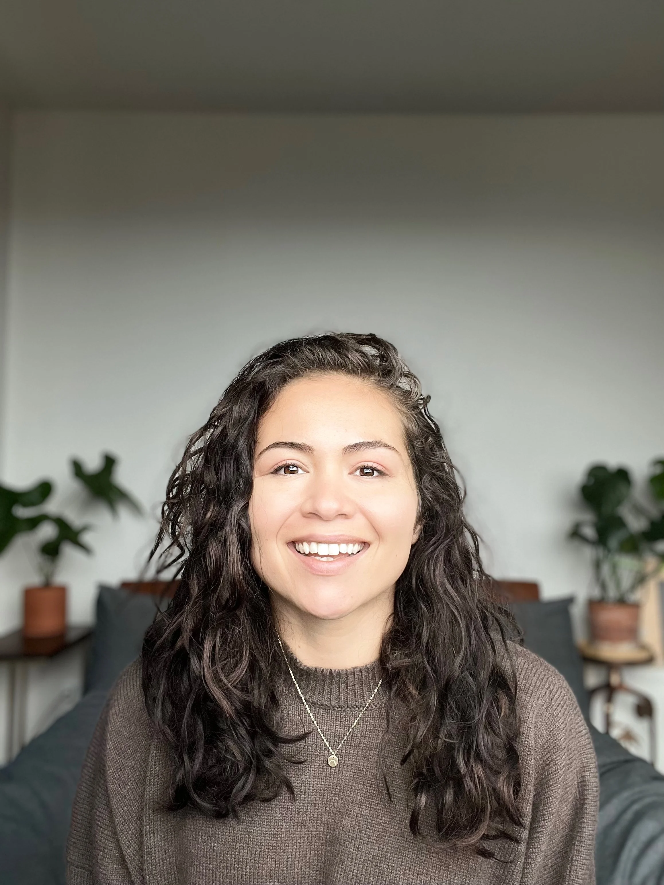 A woman with curly dark hair, smiling, wearing a brown sweater and a necklace, sitting in a room with potted plants in the background.