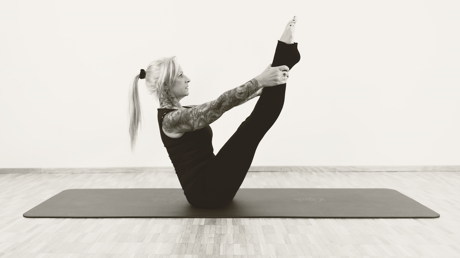 Woman with tattoos practicing yoga on a mat in a studio, sitting on her sit bones with legs extended upward.