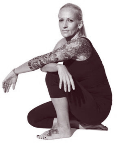 A woman with tattoos on her arm, sitting cross-legged and looking at the camera, wearing a black sleeveless top and black pants, in a black and white photo.