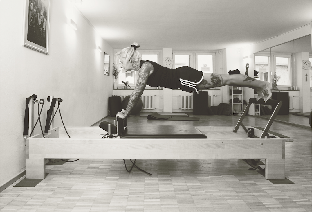 A woman with tattoos doing a plank exercise on a Pilates reformer machine in a gym room.