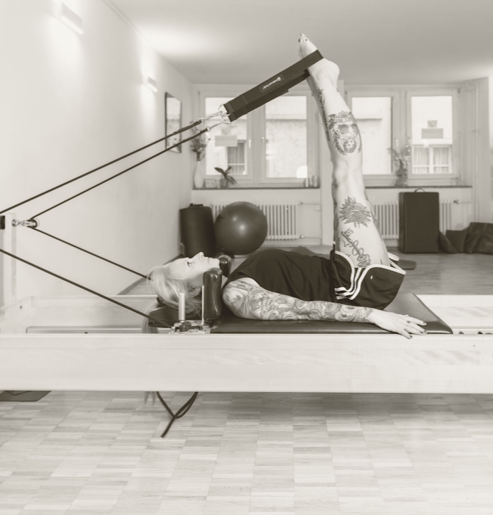 Woman with tattoos lying on a Pilates reformer machine, raising one leg with a leg extension attachment in a gym or studio.