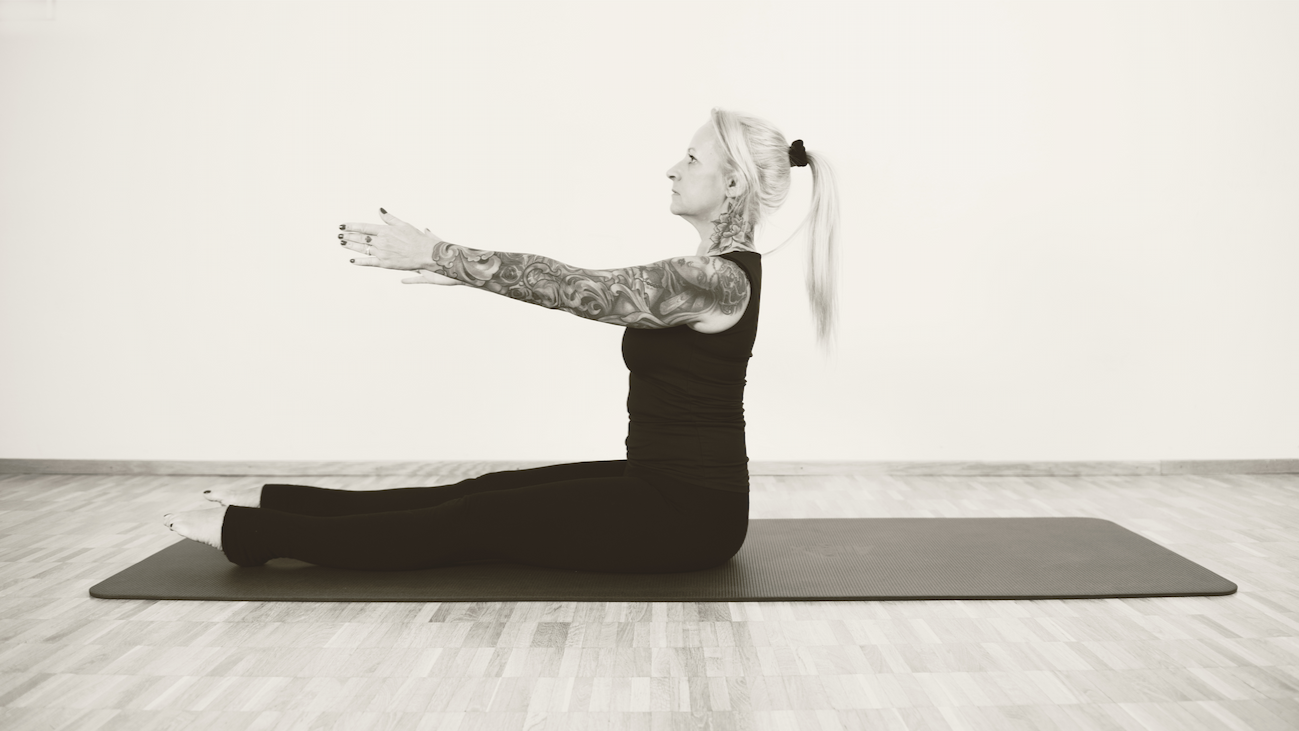 Nina Haldemann practicing Pilates indoors on a  mat, sitting with legs extended and arms stretched forward. She has tattoos on her arms and blonde hair tied back in a ponytail.