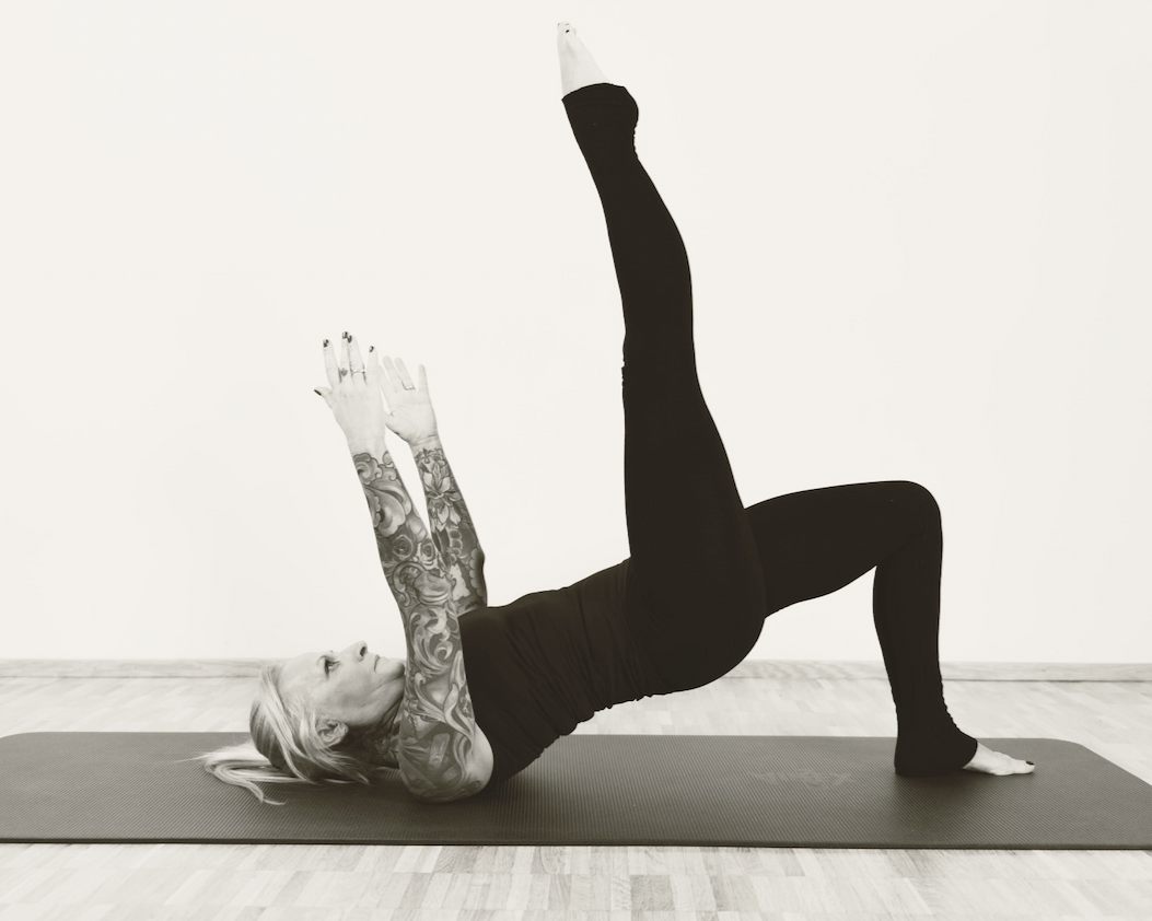 Woman with tattoos doing a Pilates spine curl with raised leg on a mat in a studio with a plain wall.