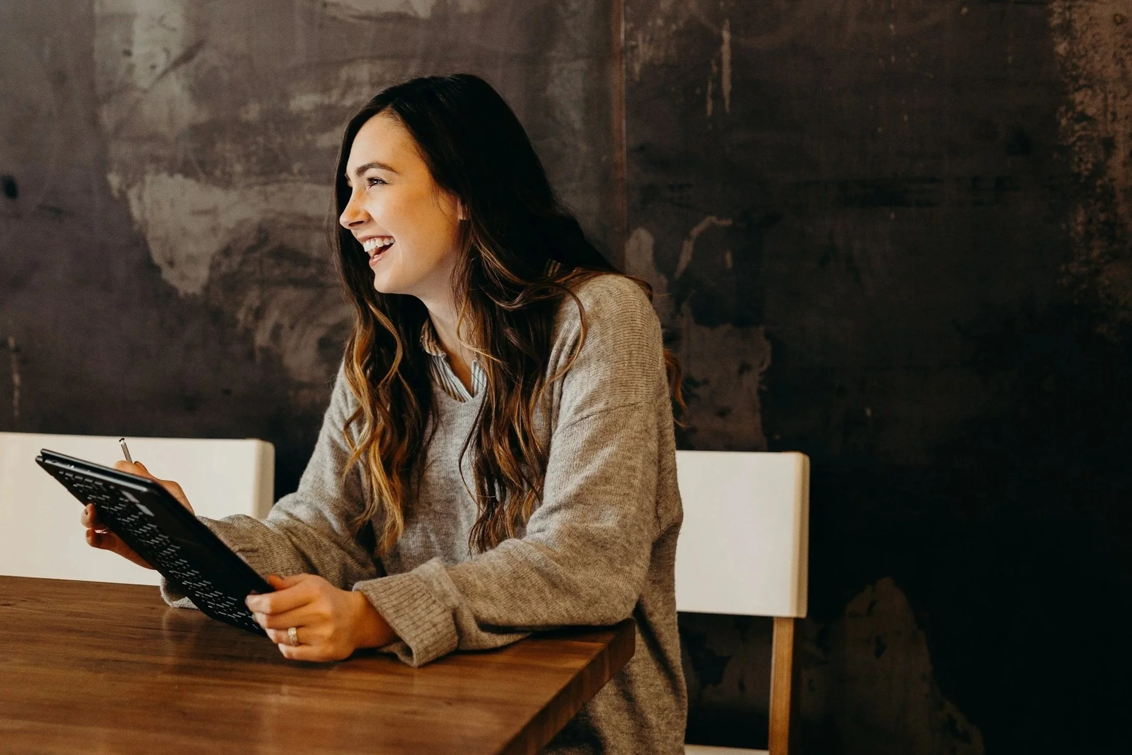 A woman with long dark hair smiling and holding a tablet while sitting at a wooden table in a dimly lit room with dark textured walls.
