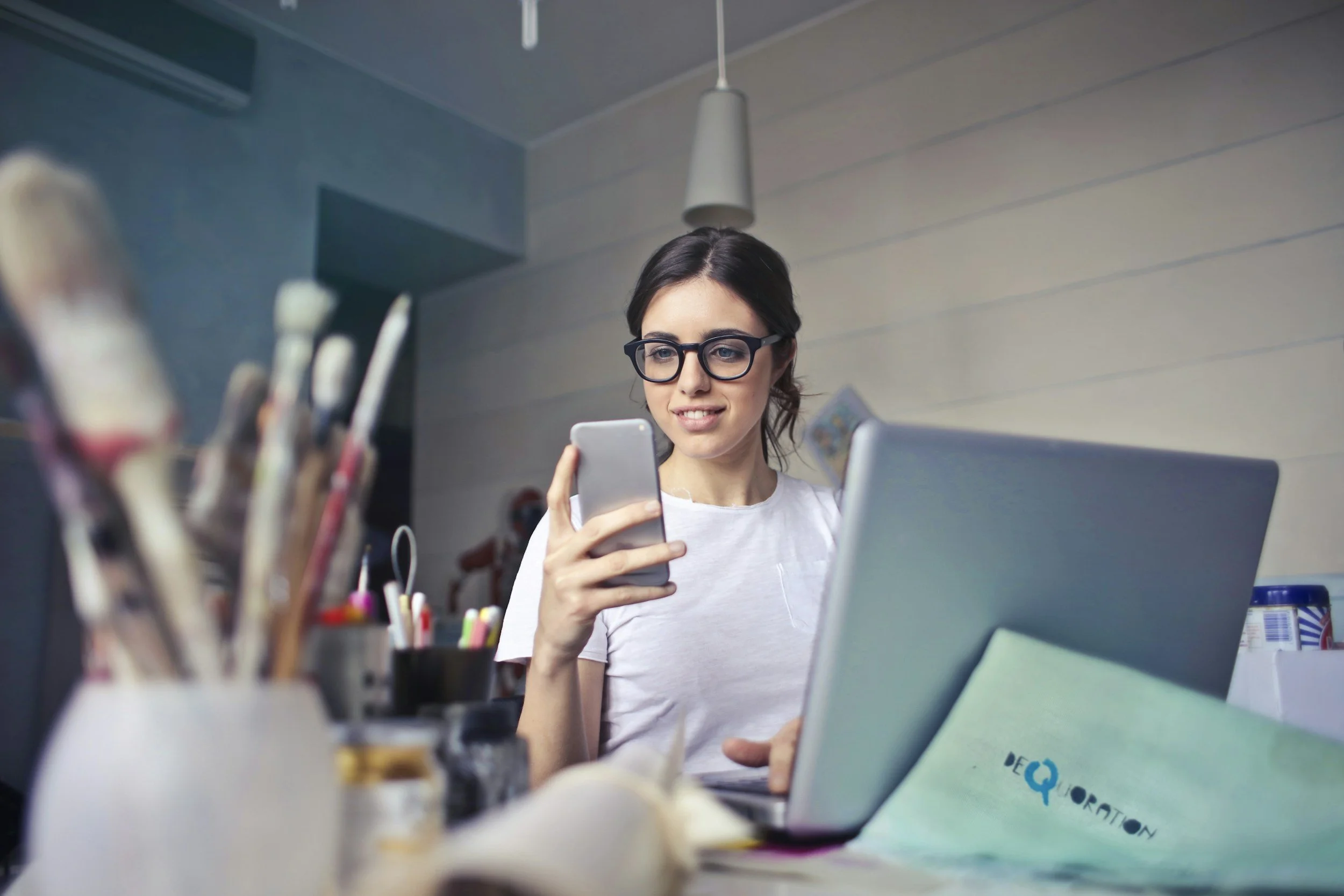 Young woman with glasses and brown hair smiling while looking at her phone in an office or studio space, with art supplies and a laptop visible on the table.