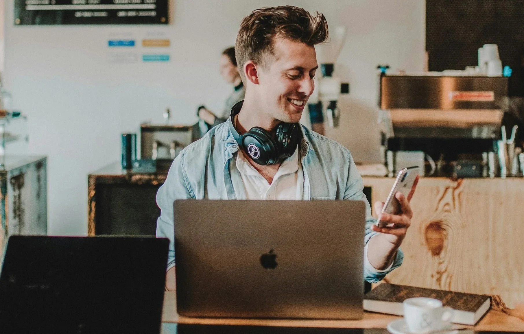 A young man with short brown hair, smiling, wearing headphones around his neck, sitting at a desk in a cafe or workspace, using his smartphone with a laptop in front of him. The background features a counter with various equipment and another person working.