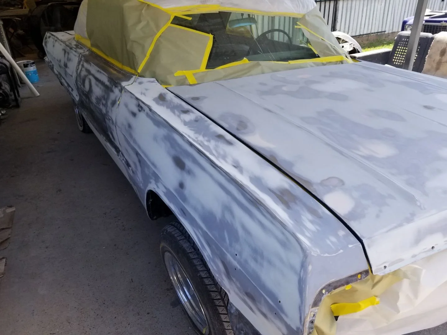 A car in the process of being repaired or restored, with its body panels sanded and primed, covered with masking paper and tape, parked in a garage or workshop.