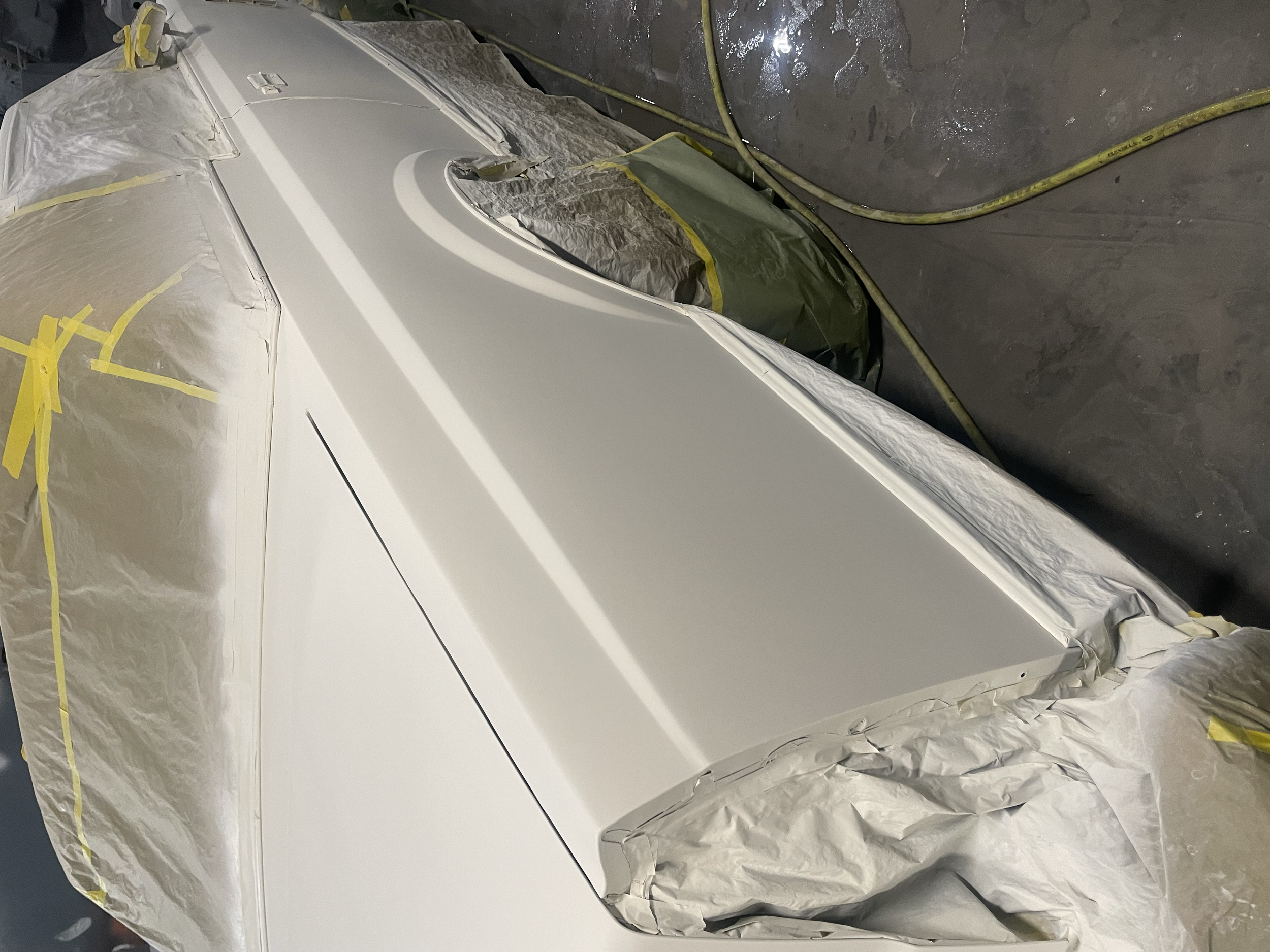A car in the midst of a paint or coating process, covered partially with protective paper and masking tape, with spray painting equipment nearby on a workshop floor.