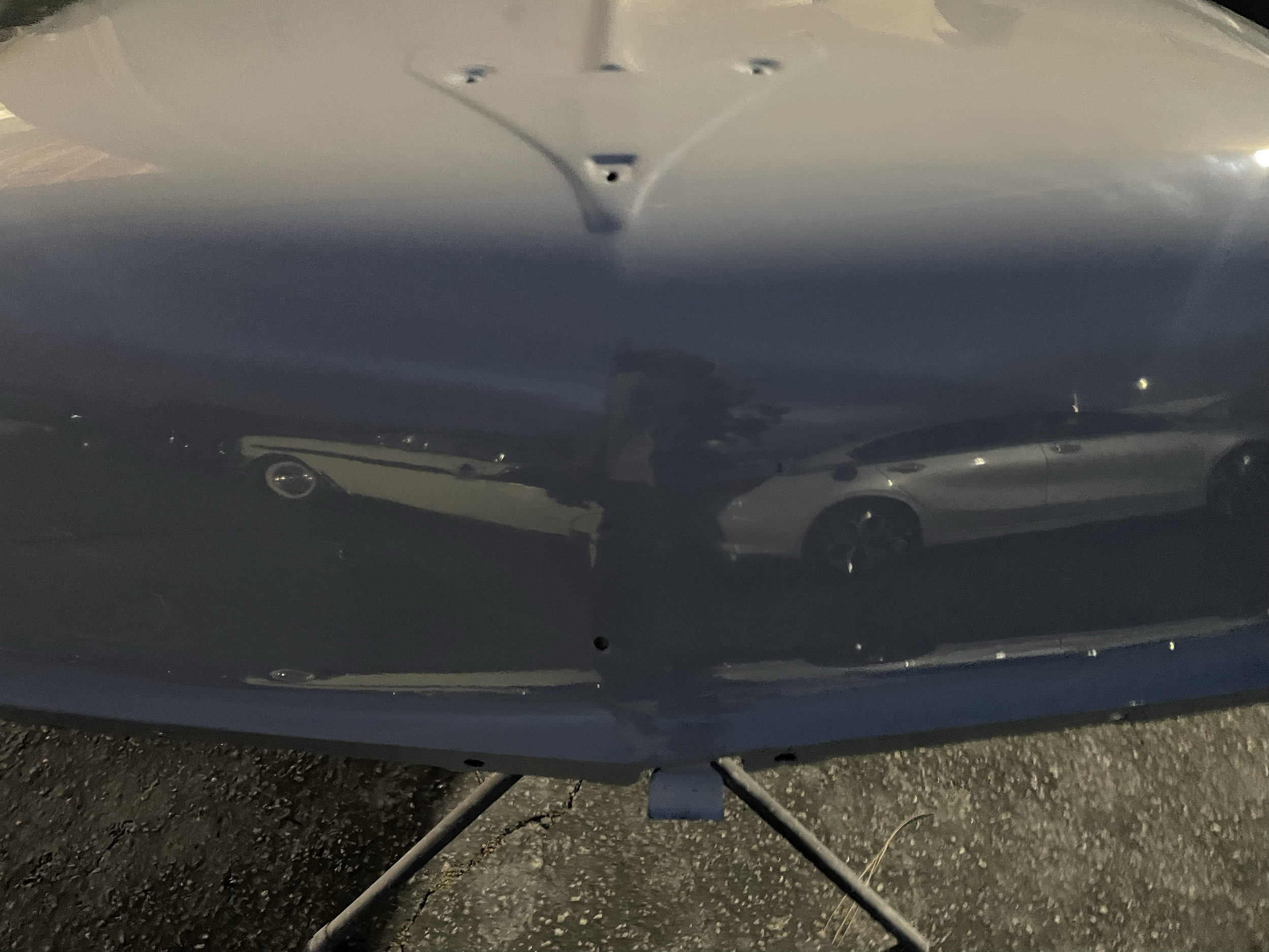 Dark-colored vehicle with a reflective surface, parked on a rough asphalt surface, with a tripod-mounted device or camera over the hood.