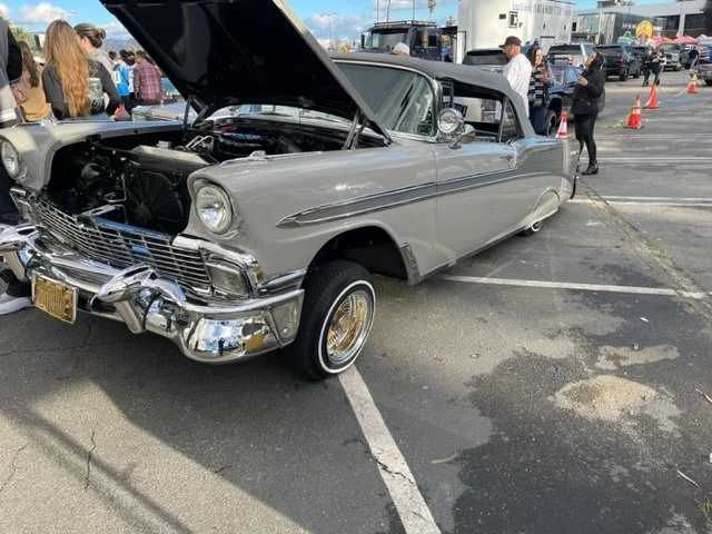 A vintage gray convertible car with its hood open at a car show, surrounded by people and traffic cones.
