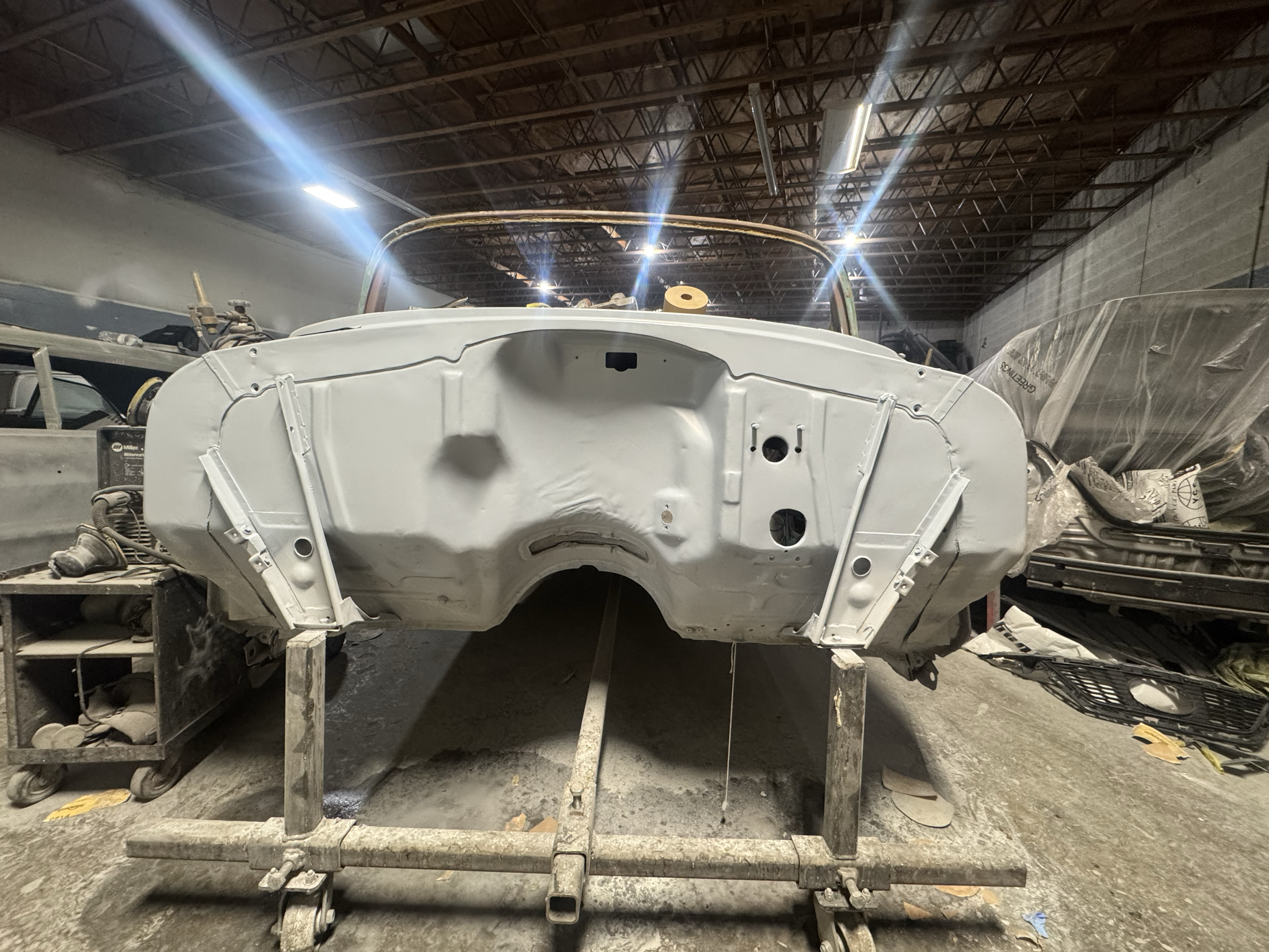 The image shows the front frame of a classic car, possibly a vintage model, on a metal stand inside a workshop or garage. The background includes a high ceiling with exposed wooden beams and ductwork, with other vehicle parts and equipment around.