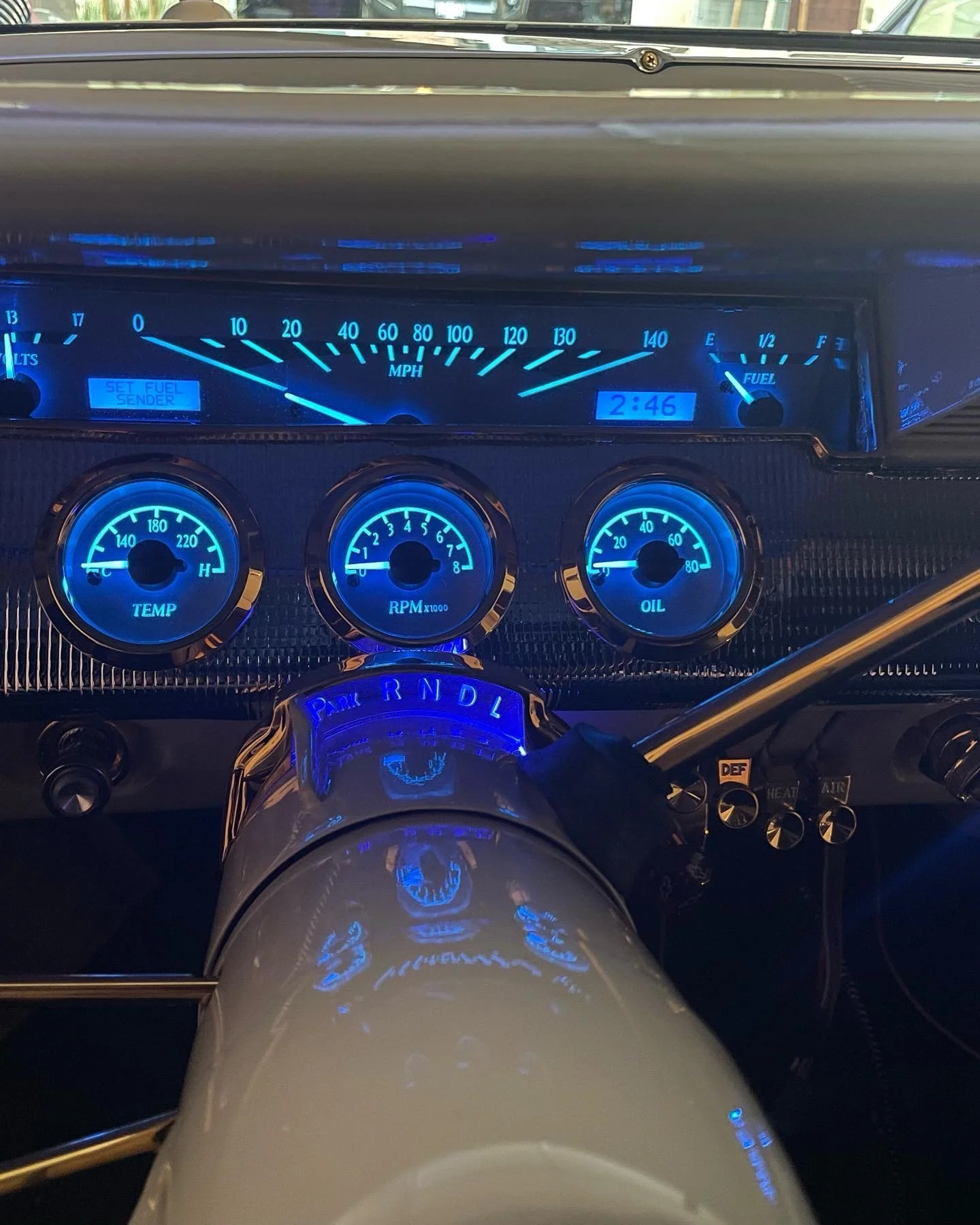 Close-up of a vintage car dashboard illuminated with blue lights, showing a speedometer, RPM gauge, oil gauge, fuel gauge, and a digital clock reading 2:46, with a steering wheel in the foreground.