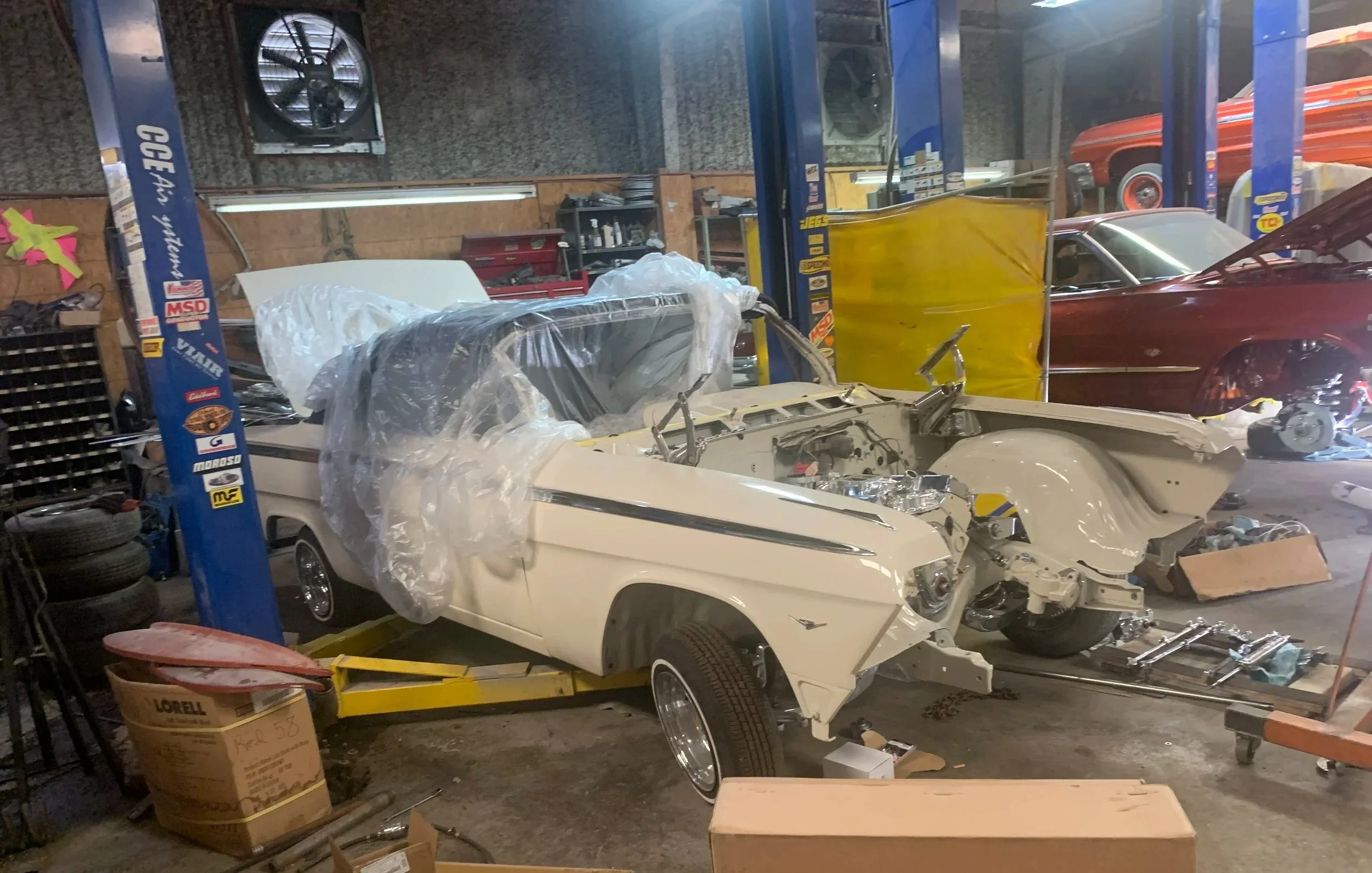 A garage workshop with vintage cars, including a partially disassembled white car in the foreground and a red car in the background, along with tools, tires, and miscellaneous auto parts.