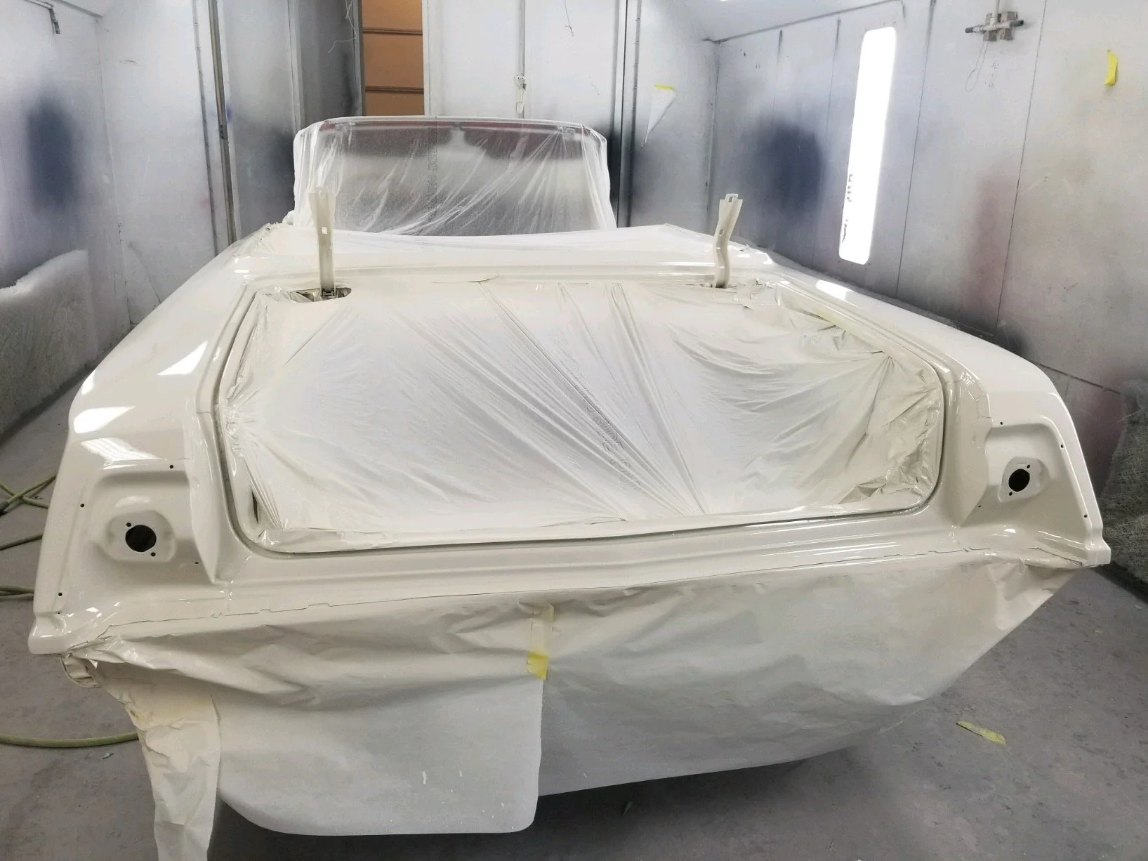 A car is covered with white protective plastic, with areas taped off for painting or repair in a workshop or spray booth.