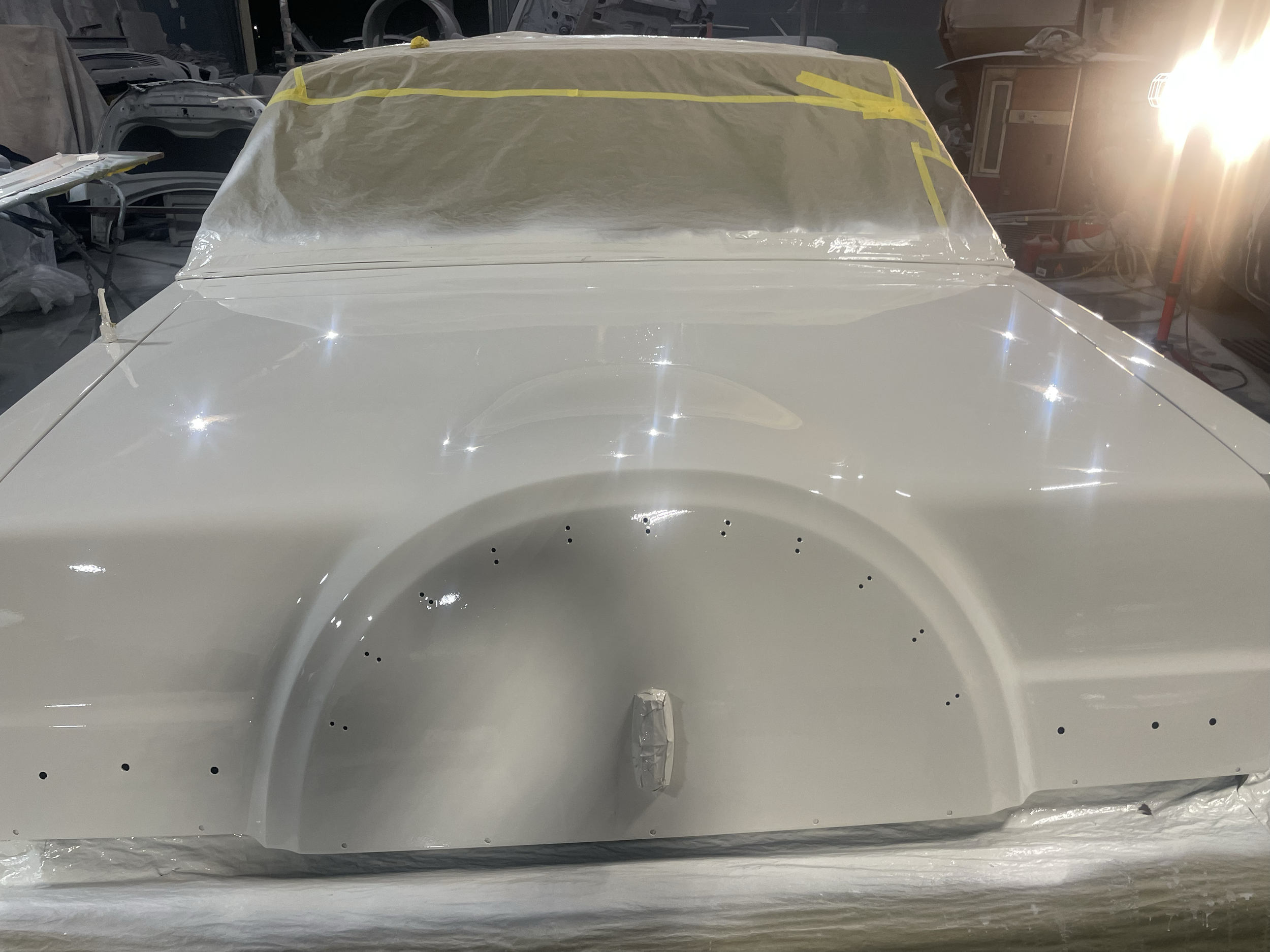 A white classic car undergoing restoration, with parts covered in protective paper and yellow painter's tape, inside a workshop illuminated by bright lights.