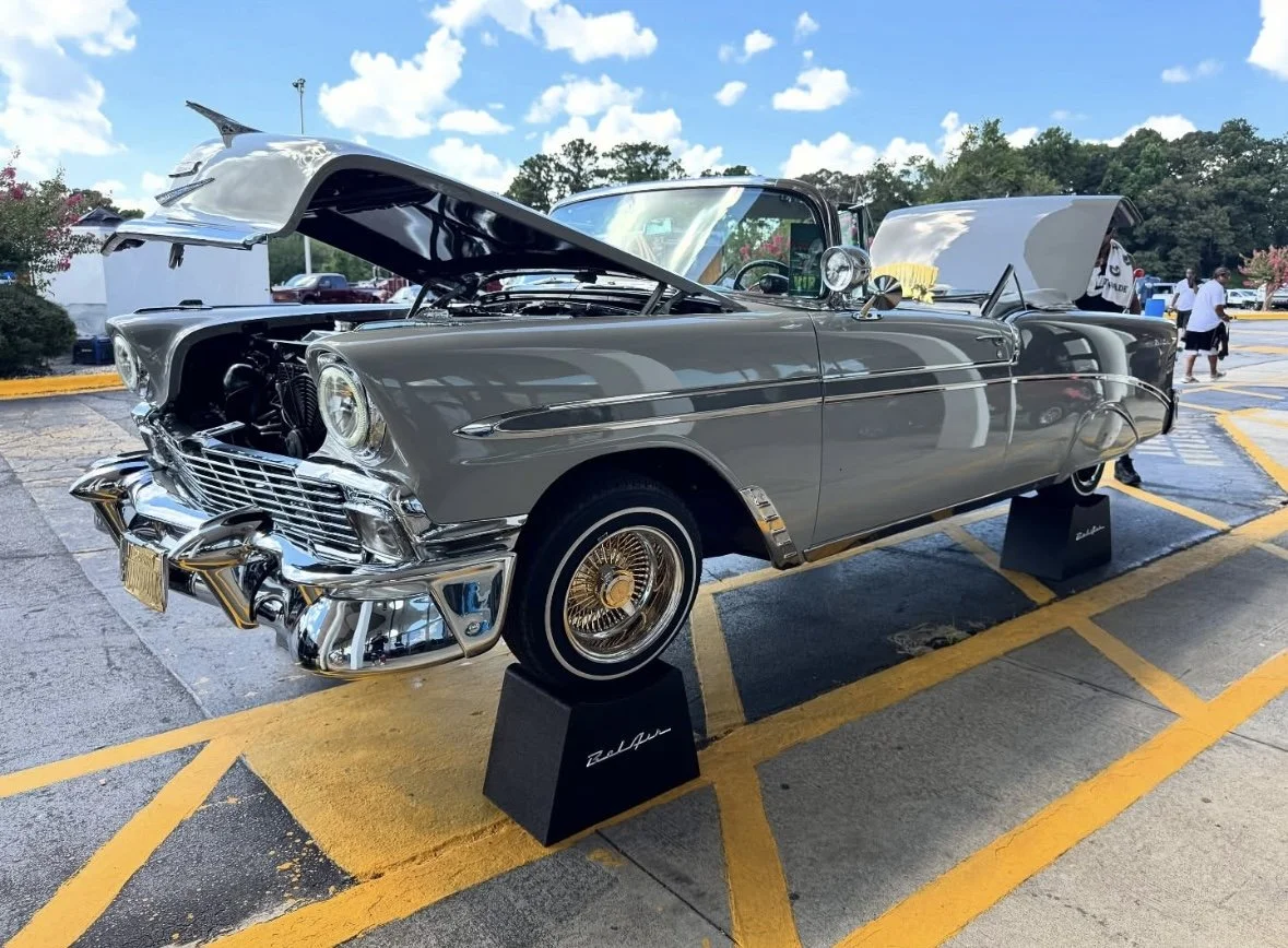 A vintage silver convertible car on display at an outdoor car show, with its hood open revealing the engine.
