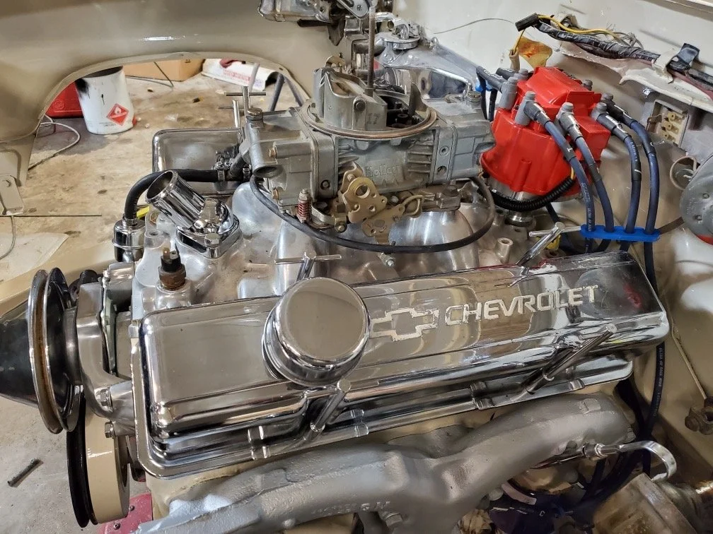A Chevrolet car engine with chrome valve covers, red distributor cap, and various wires and parts, in a workshop setting.