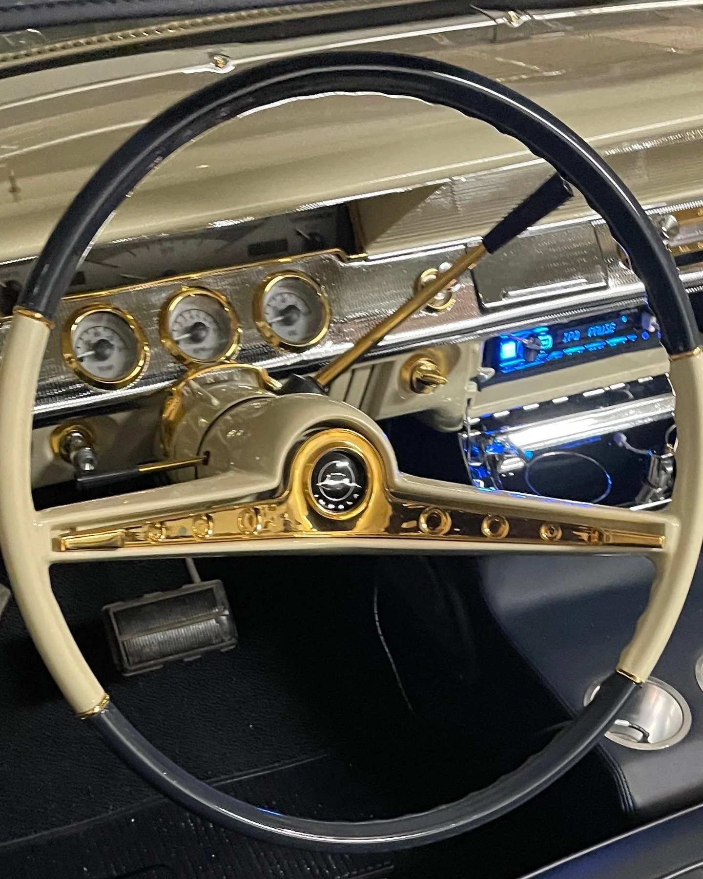 Close-up of a vintage car dashboard and steering wheel with gauges, gold accents, and a blue digital display.