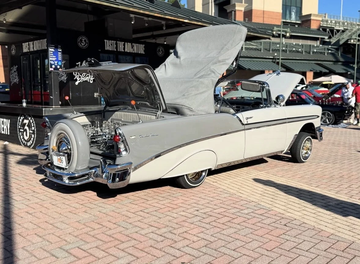 A vintage two-tone white and gray convertible car with its hood open, parked at a classic car show on a brick-paved area.