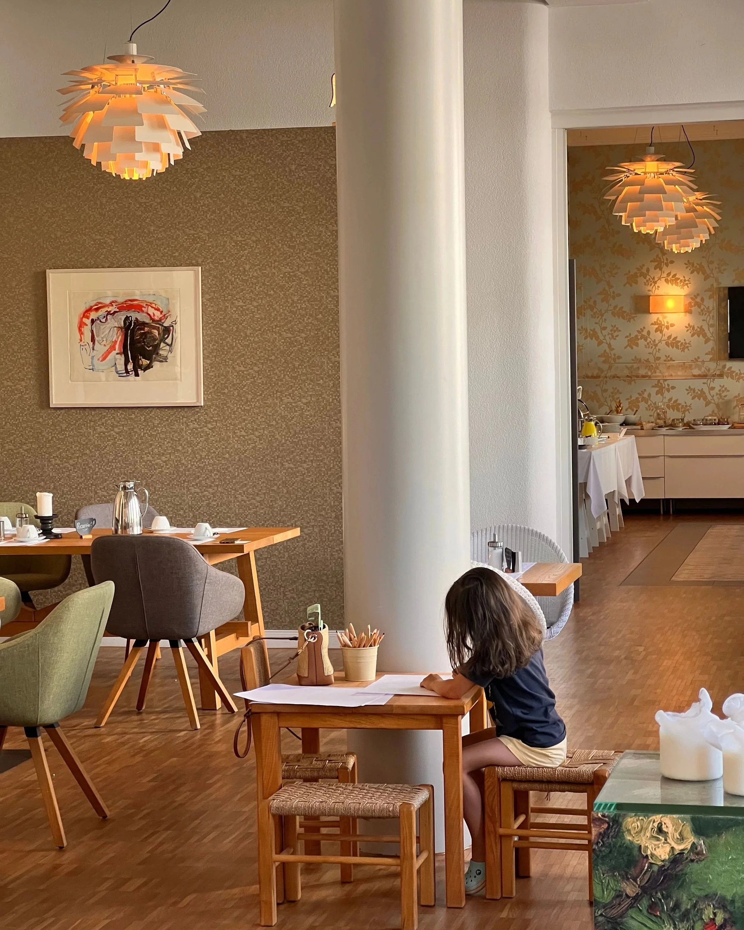 Une pièce de restaurant ou de salle à manger avec des lampes modernes, une peinture abstraite, plusieurs tables et chaises, un enfant assis à une table, bien accueilli, et un décor chaleureux. Kid friendly, adapter aux enfants et bébés