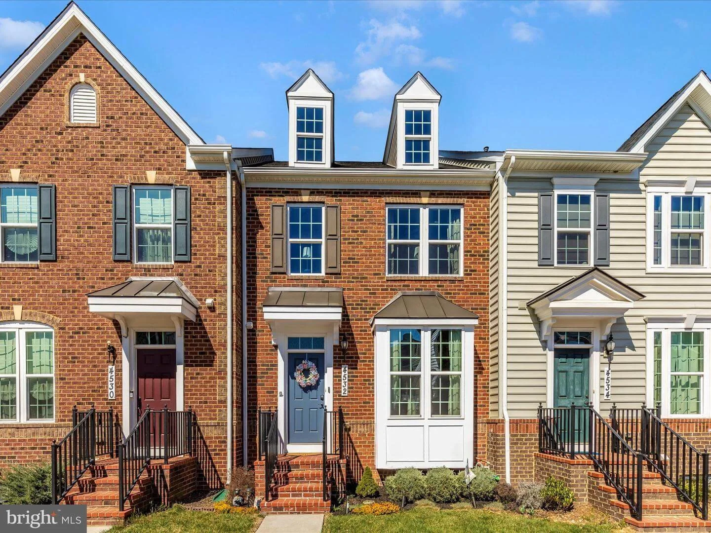 Three attached townhouses with brick and siding exteriors, three stories, steep roofing with dormer windows, front porches with stairs, and small front lawns.