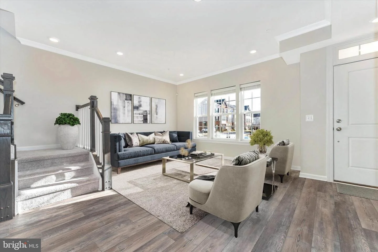 Living room with a grey couch, two beige armchairs, a glass coffee table, and decor on the wall, illuminated by natural light from multiple windows.