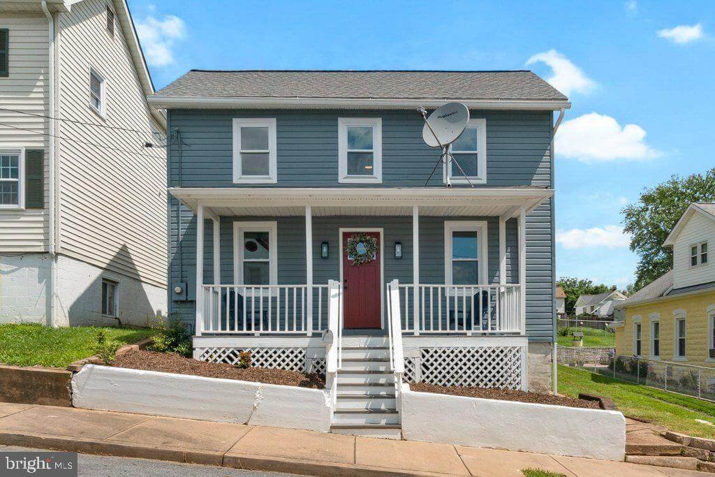 A two-story house with blue siding, white trim, and a front porch with white railings. The house has a red front door with a wreath, four windows on the front, and a satellite dish on the roof. There are stairs leading up to the porch, and the yard i