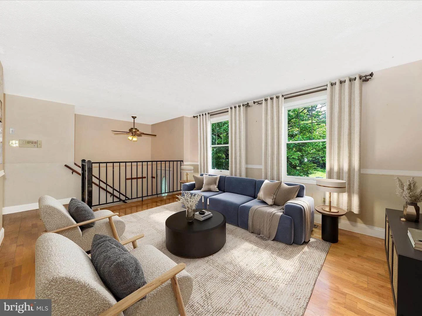 Living room with beige walls, hardwood floors, large window with white curtains, blue sofa with pillows, round black coffee table, twin white armchairs with gray pillows, side table with lamp, and a black console table with decoration, overlooking a 