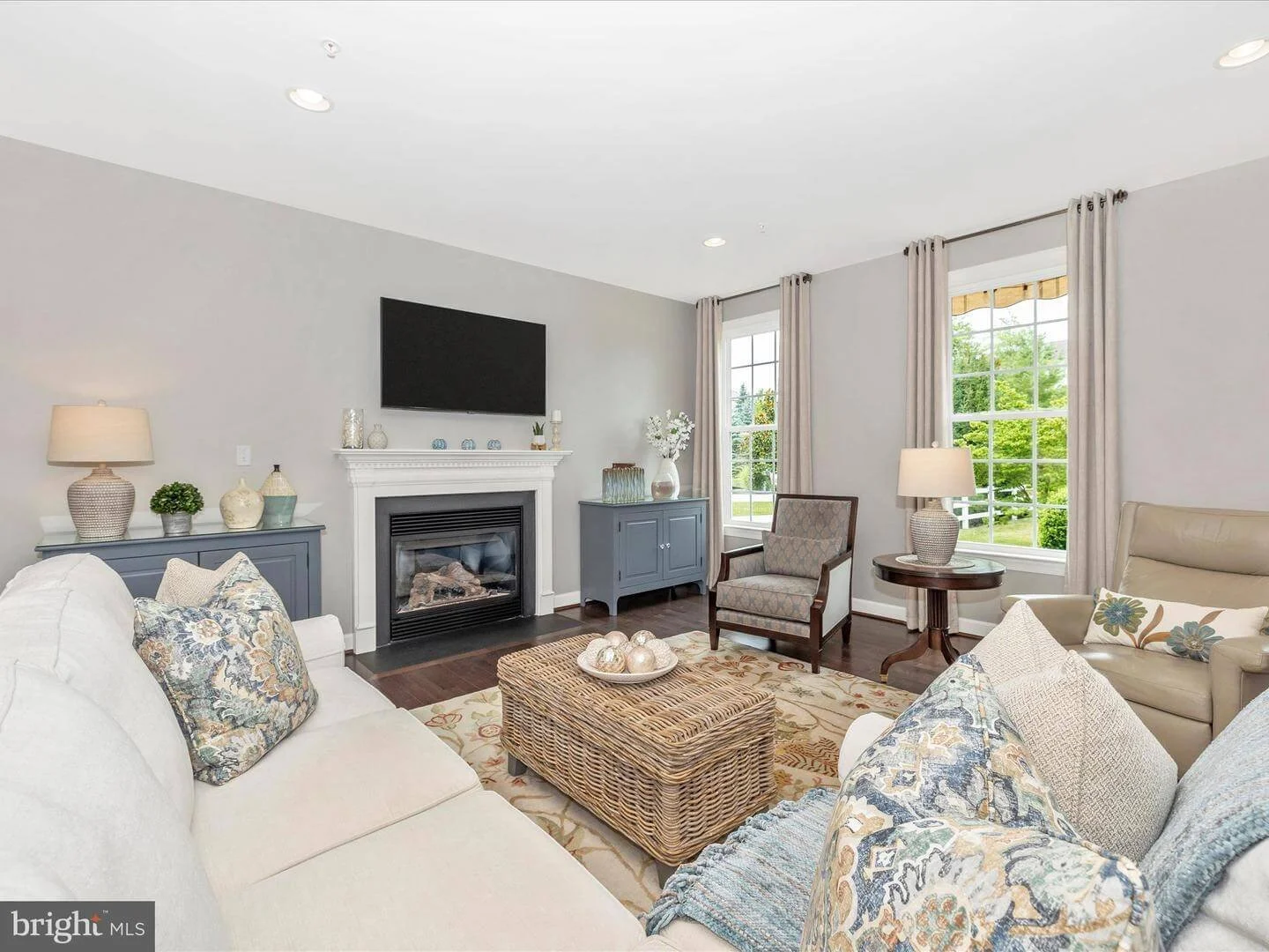 Living room with white sofa, beige armchair, wicker coffee table, and side tables with table lamps. Fireplace with decorative items on mantel, flat-screen TV above. Two large windows with beige curtains showing green outdoor scenery.