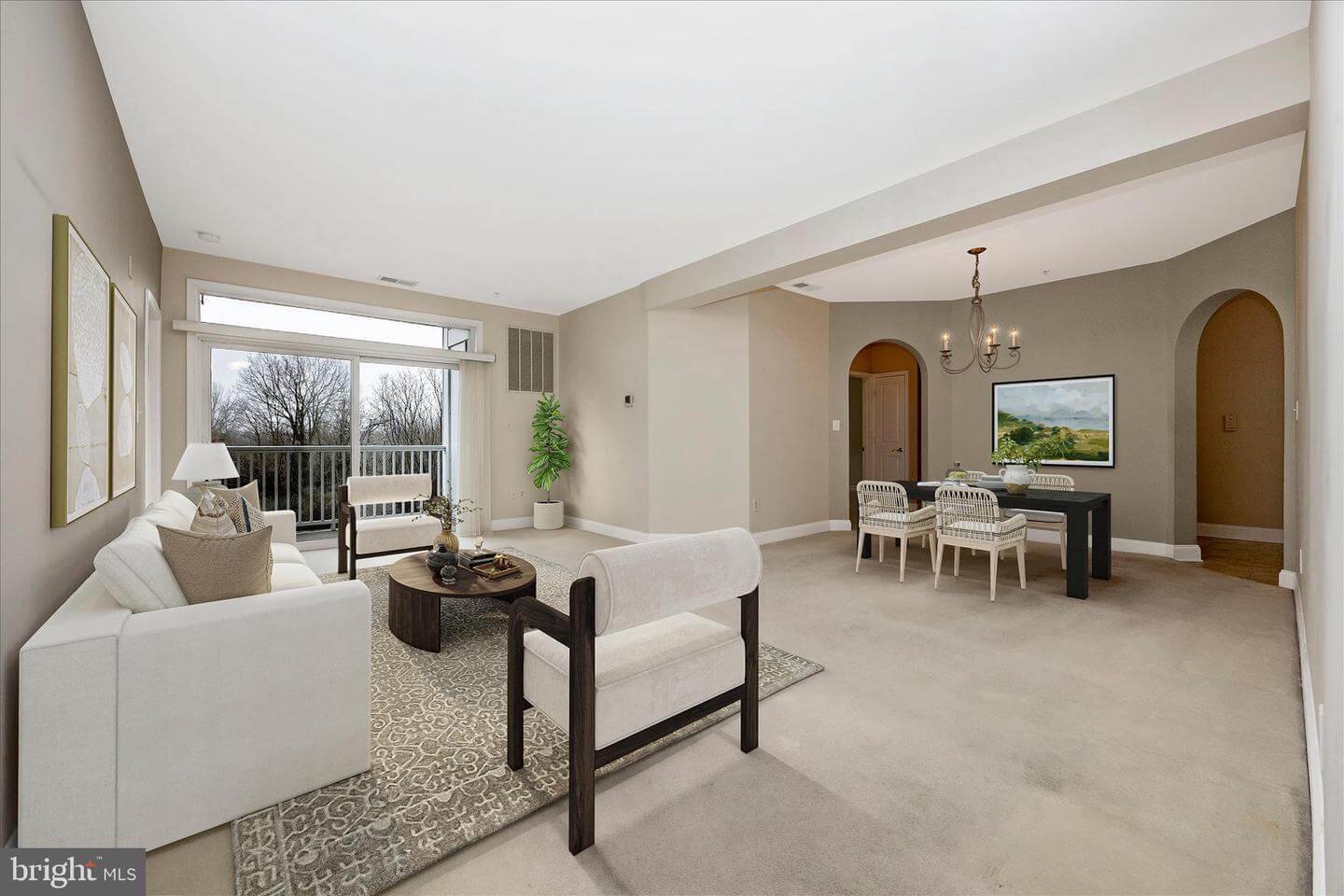 Spacious living room with white sofa, beige armchair, round coffee table, and large window with balcony. Adjacent dining area with black table, white chairs, and a wall-mounted TV. Neutral-colored walls and carpet, with a chandelier hanging above the