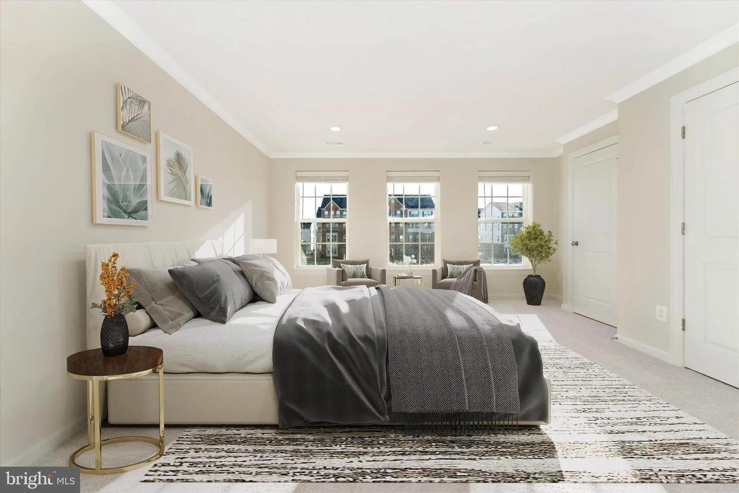 Bright bedroom with a large bed, neutral-colored bedding, and wall art above, with windows at the far end, a potted plant, and a patterned rug.