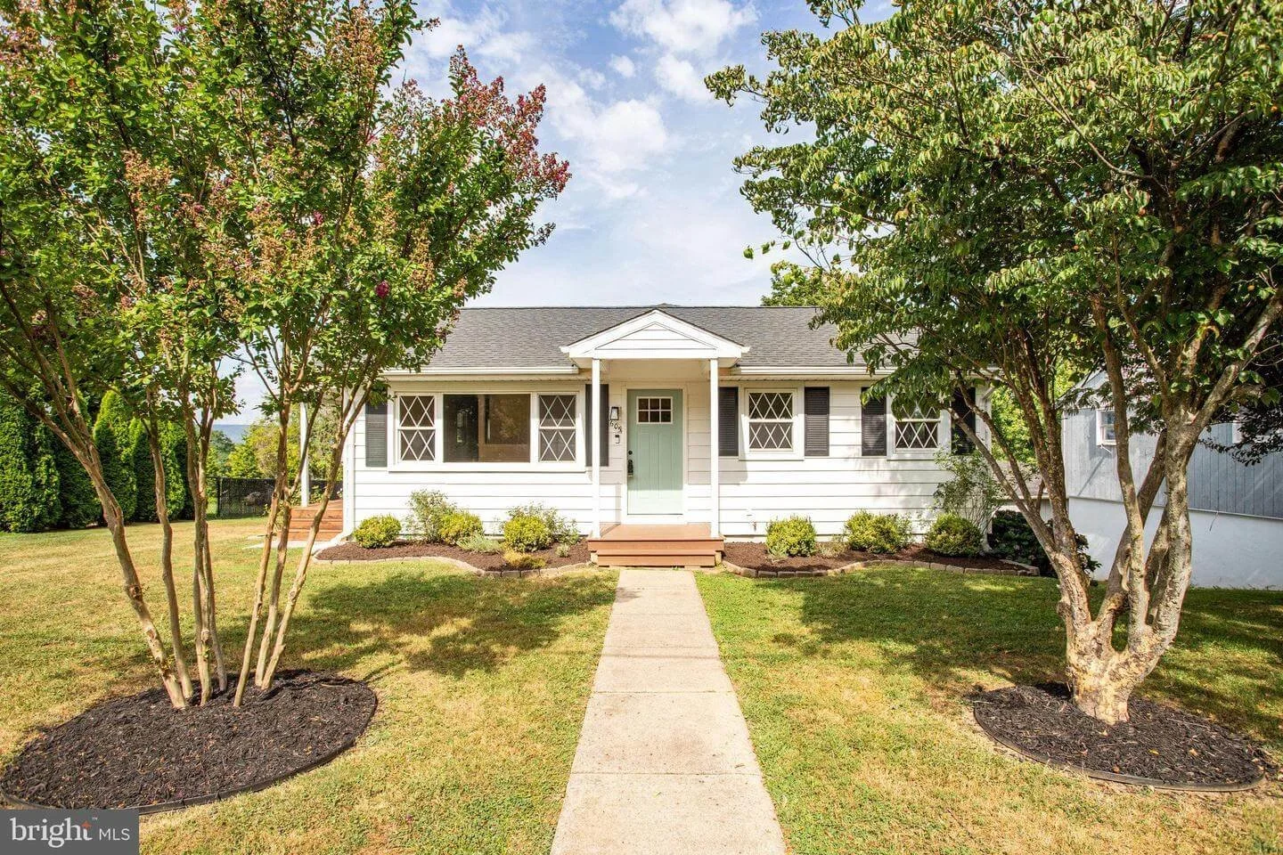 A white single-story house with black shutters and a light green front door, surrounded by a well-maintained lawn with two small trees and bushes, under a partly cloudy sky.