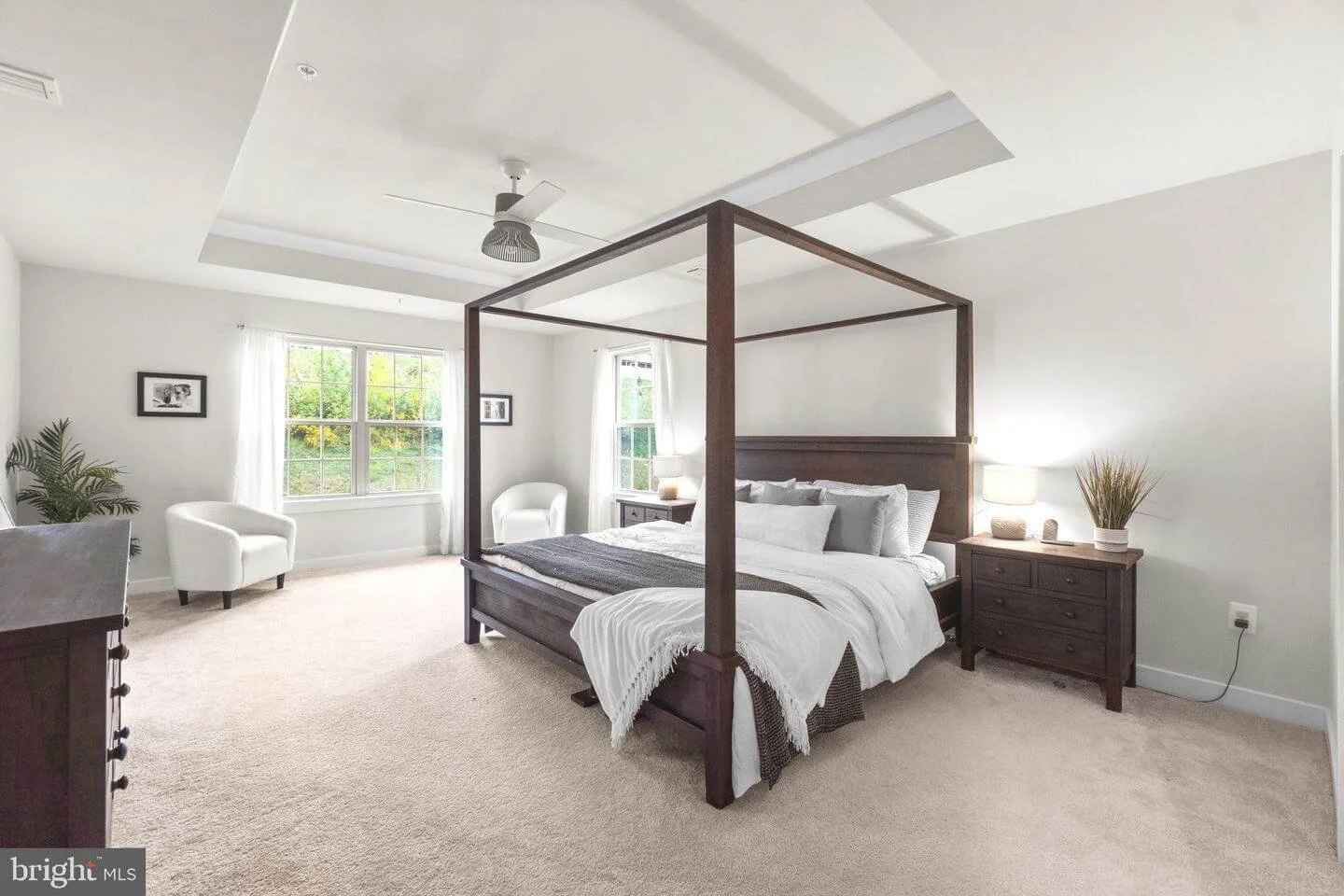 Bright bedroom with a four-poster bed, white bedding, nightstands, lamps, a ceiling fan, and sitting area with chairs near large windows showing green trees outside.