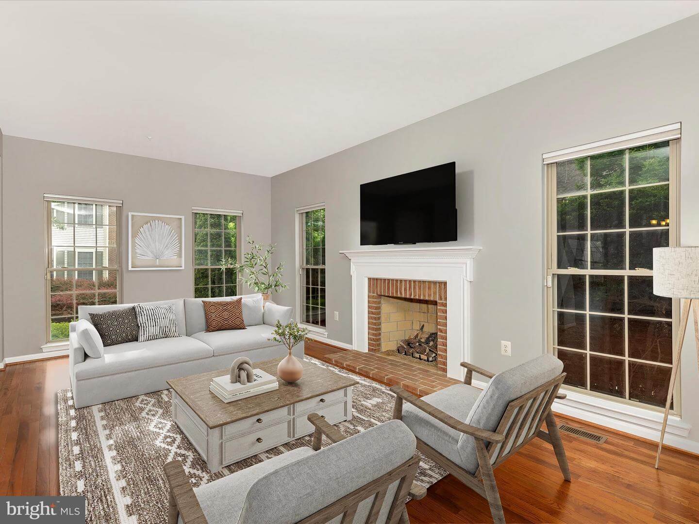 Living room with gray walls, three large windows, white sofa with decorative pillows, two gray armchairs, a wooden coffee table with decorations, brick fireplace with white mantel, wall-mounted TV above fireplace, a patterned rug, and a floor lamp.
