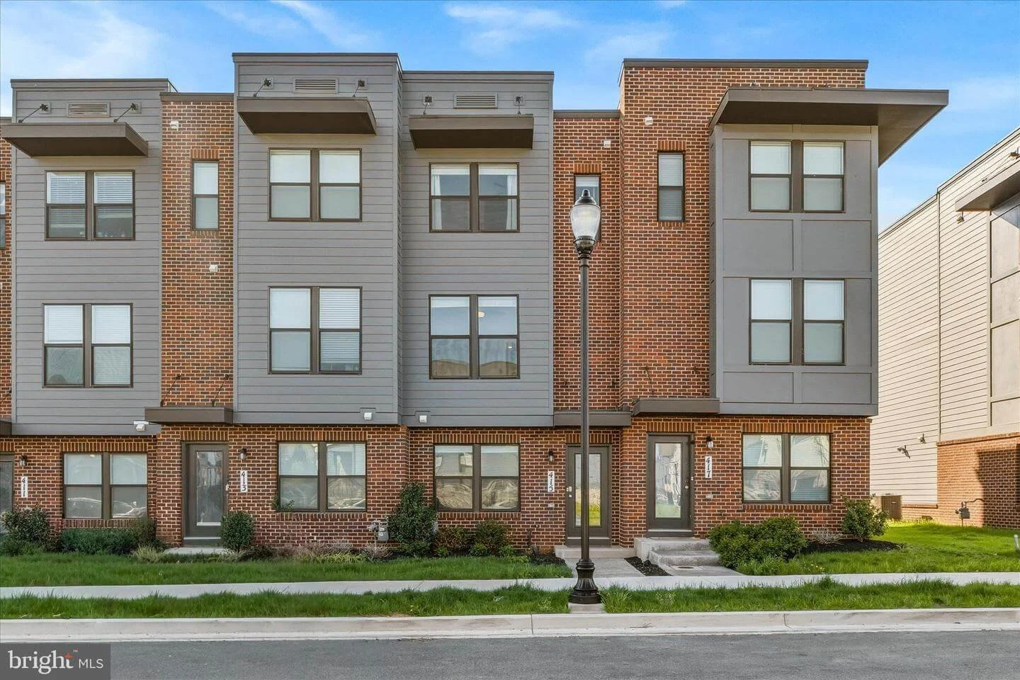 Multi-story residential building with brick and gray siding, with a streetlamp in front, green lawn, and steps leading to doors.