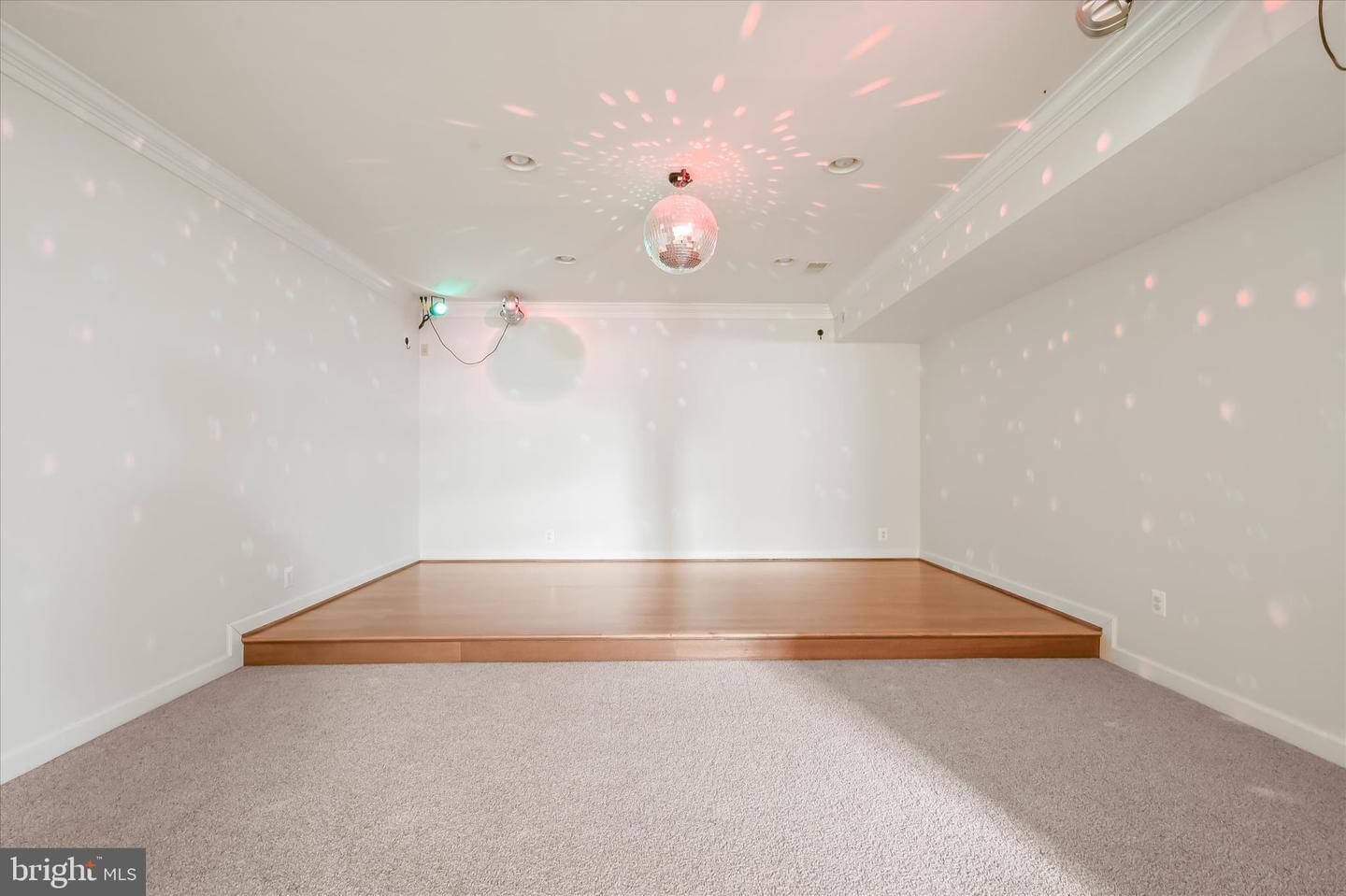 Empty room with white walls, a light-colored carpet, a small wooden stage area, and a disco ball on the ceiling casting colorful lights.