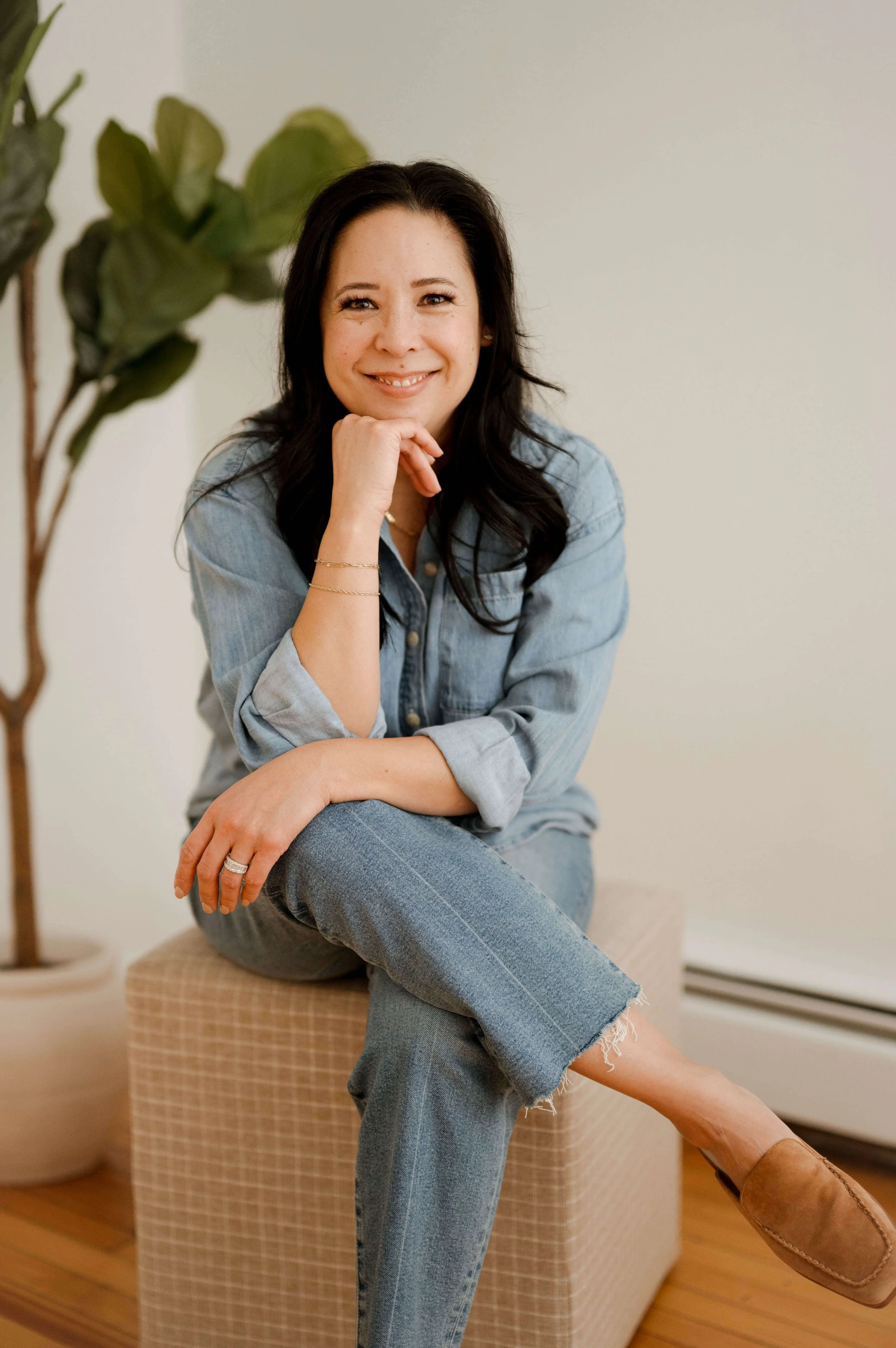 A woman with dark hair sitting on a beige ottoman, wearing a denim jacket and ripped jeans, smiling and resting her chin on her hand, with a green plant and a beige checkered ottoman in the background.