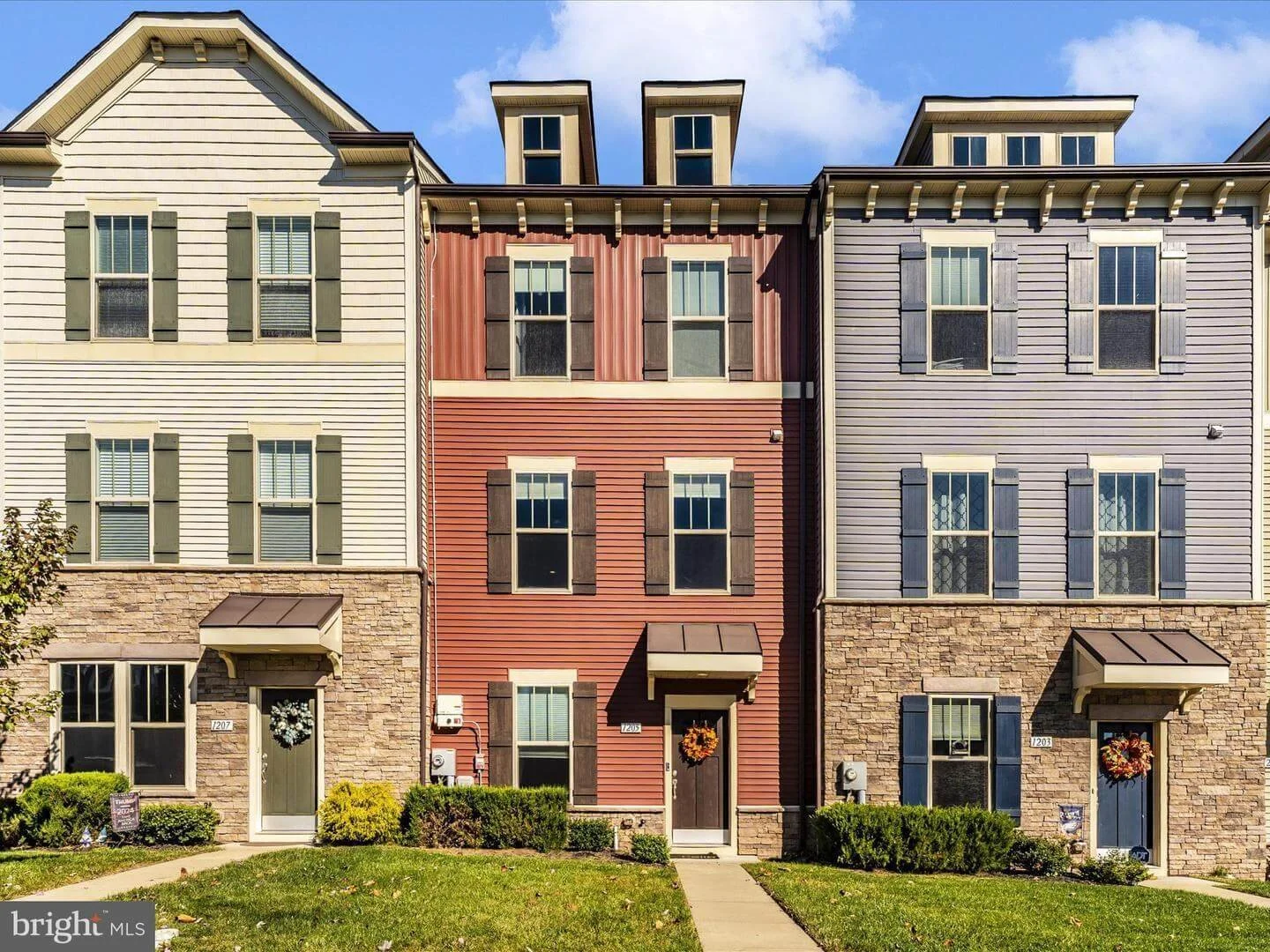 Three connected multi-story townhouses with colorful exteriors, stone lower walls, and front doors decorated with wreaths, set against a blue sky.
