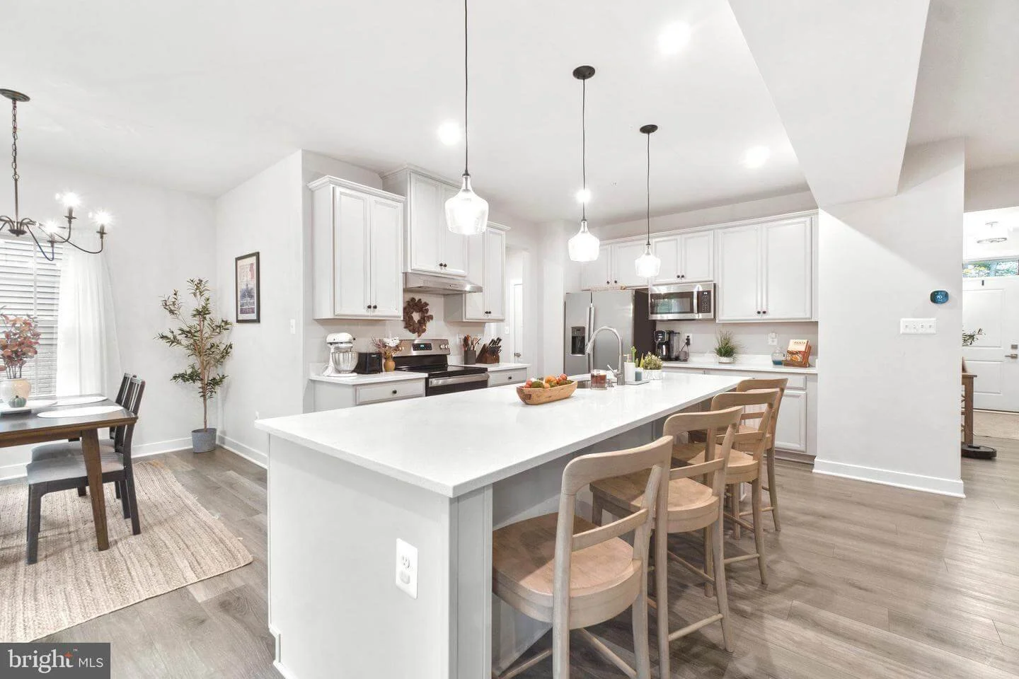 Modern white kitchen with island, wooden stools, stainless steel appliances, and natural light.