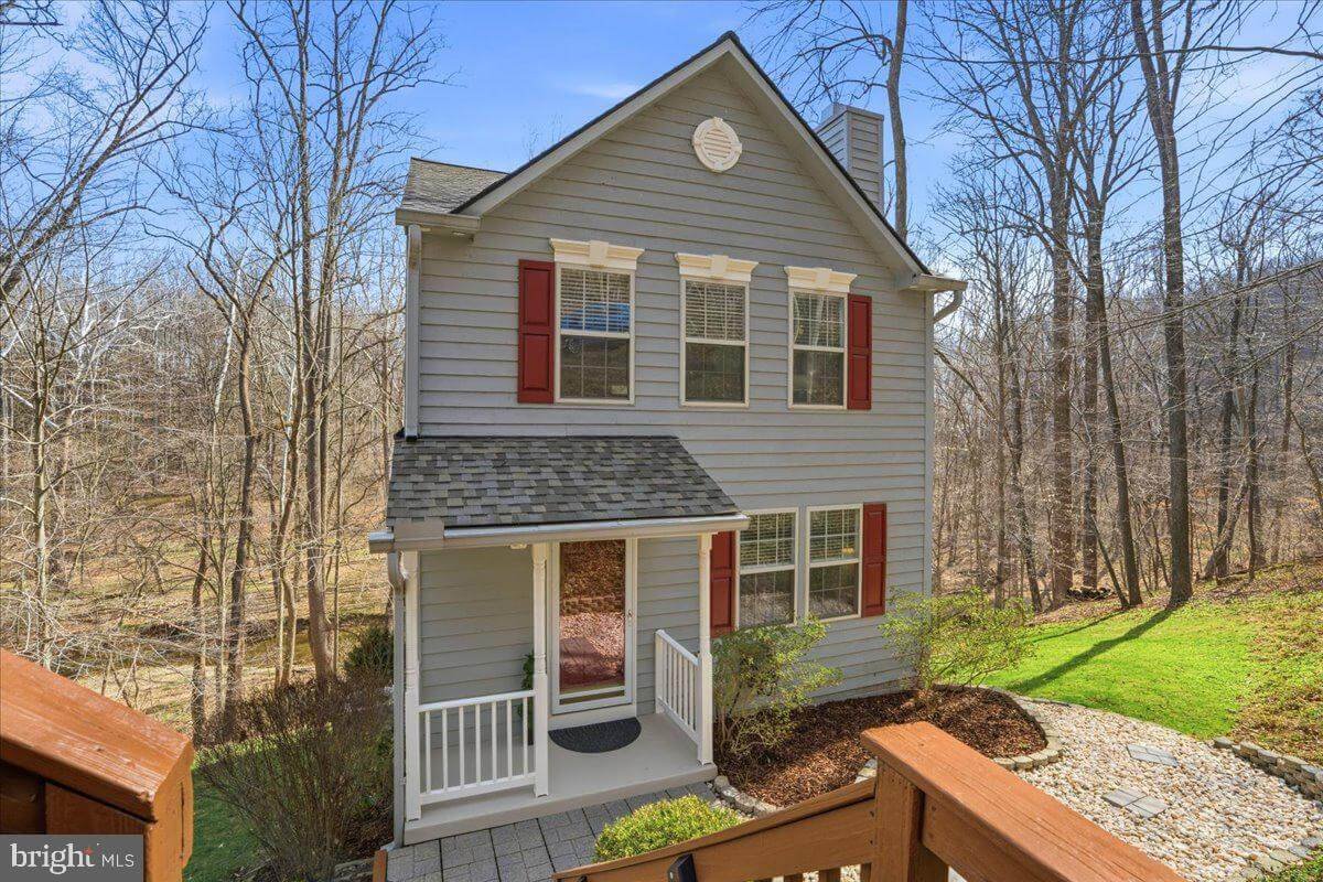 A two-story house with gray siding, white trim, red shutters, and a gabled roof. It has a front porch with white railings and a screened door. The yard has trees and a rock-lined walkway.