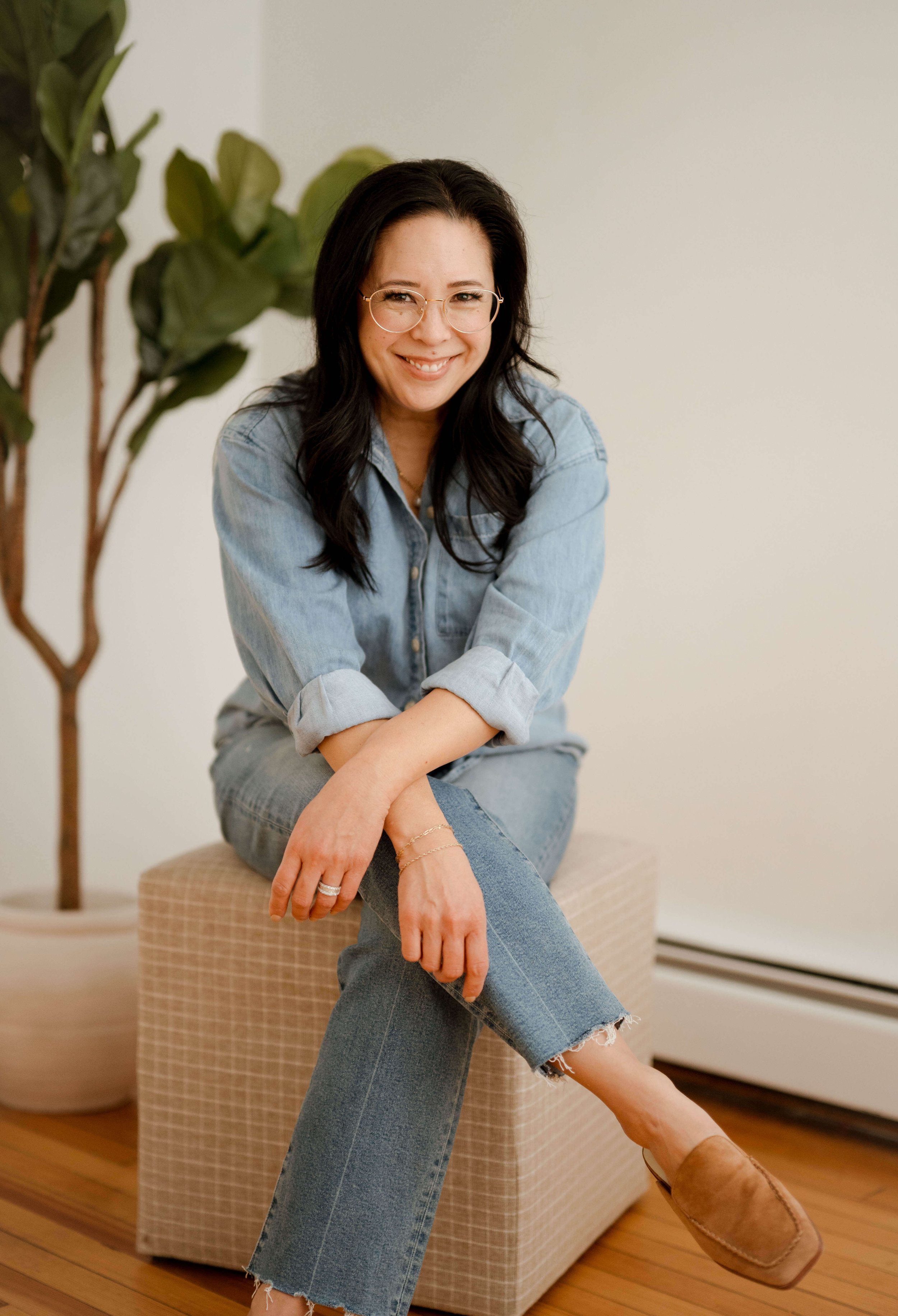 A woman with long dark hair, glasses, and light denim outfit sitting on a beige ottoman in a room with wooden floors and a green plant nearby, smiling at the camera.