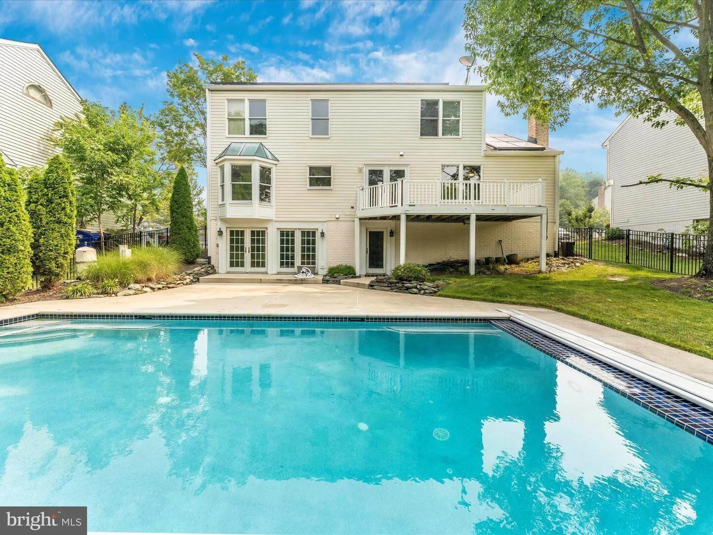 Backyard with swimming pool, patio, and white house with deck, surrounded by trees and greenery.