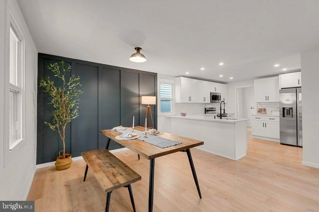 Open kitchen and dining area with a wooden table, bench, a potted plant, white cabinets, stainless steel appliances, and a dark feature wall.
