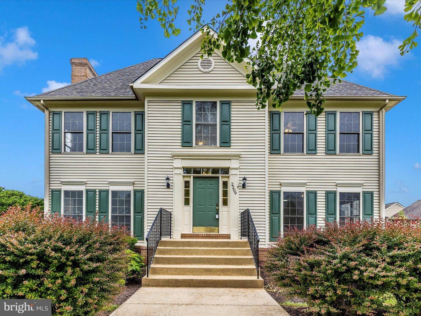 A two-story house with beige vinyl siding, green shutters, and a green front door, surrounded by greenery and shrubs under a blue sky.