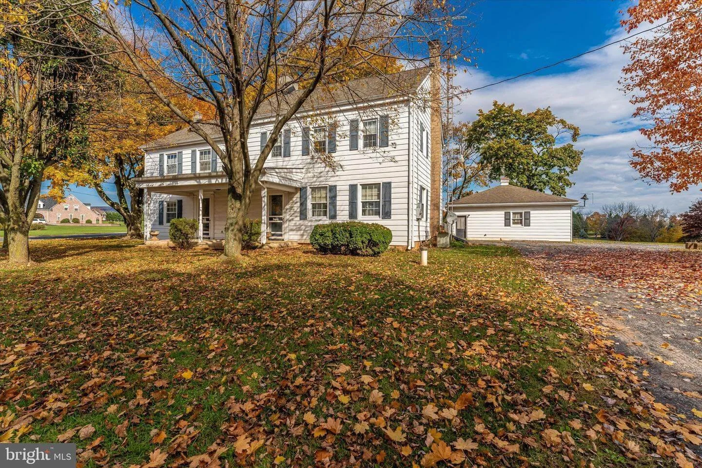 A white two-story house with black shutters, a porch, and a side extension, surrounded by trees with fallen leaves on the grassy ground and a gravel driveway.