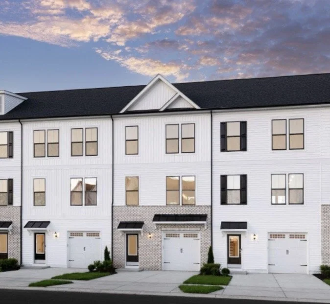 A row of modern three-story townhouses with white siding, black accents, and attached garages, under a partly cloudy sky at sunset.